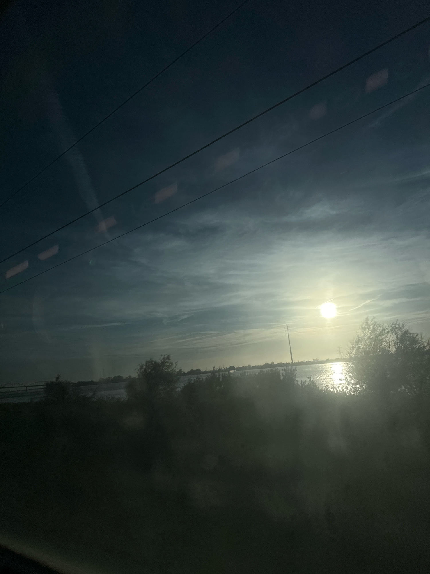 This image depicts a view of the Tagus River at sunset in Vila Franca de Xira, Portugal. The sun is low on the horizon, casting a bright glare across the water and illuminating the clouds with a soft, warm light. On the far bank of the river, a line of trees and vegetation is visible, with some power lines stretching across the sky. The foreground of the image is dark and blurred, suggesting it was taken from inside a moving vehicle, possibly a train or car, looking out through a window. The overall impression is one of a tranquil, natural landscape bathed in the fading light of day.