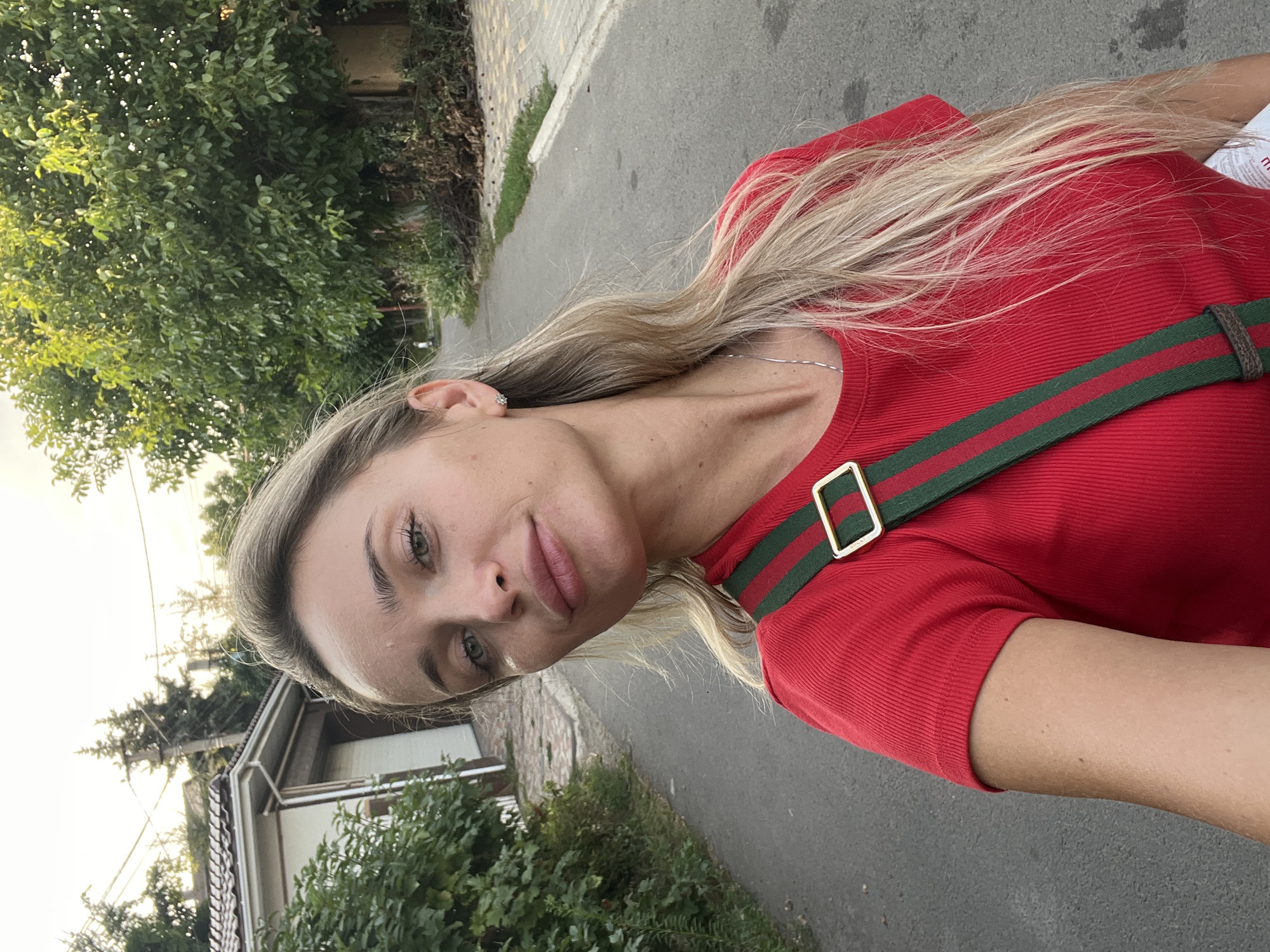 A blonde woman stands on a paved road with trees and a house in the background. She is wearing a red t-shirt with a green and red striped strap across her chest. Her hair is long and blonde, and she is looking at the camera. The sky is overcast. The location is in Fontanka, Ukraine.