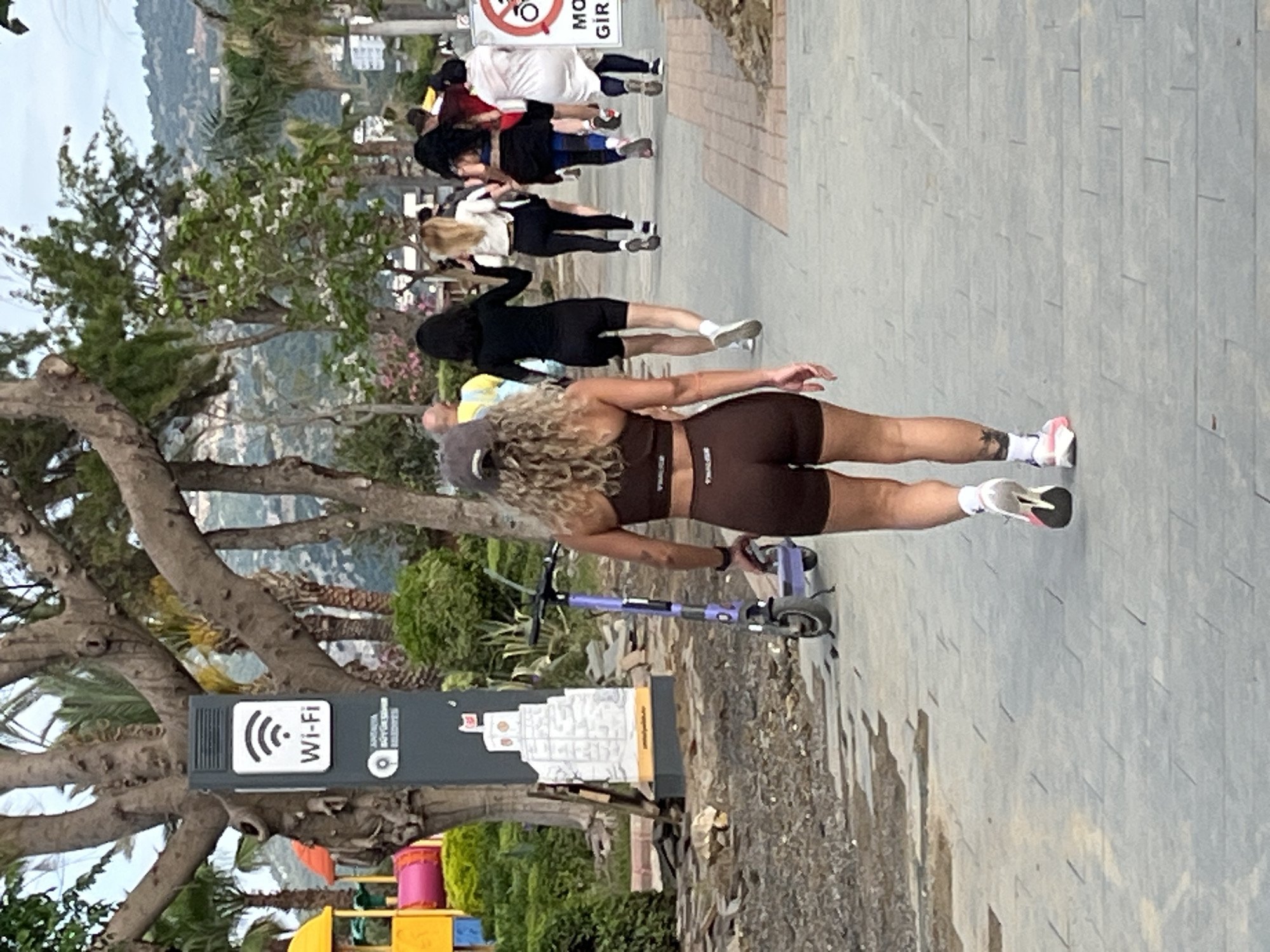 The image captures a group of individuals on a pedestrian walkway in Alanya, Turkey. A woman in brown athletic wear is standing with a purple scooter near a Wi-Fi sign and a tourism sign showing a white building, indicating an area with public internet access. Behind her, a line of people is moving along the walkway. The environment includes trees, and the background reveals a glimpse of the city and hills. The walkway is paved, and there is vegetation along its edge.