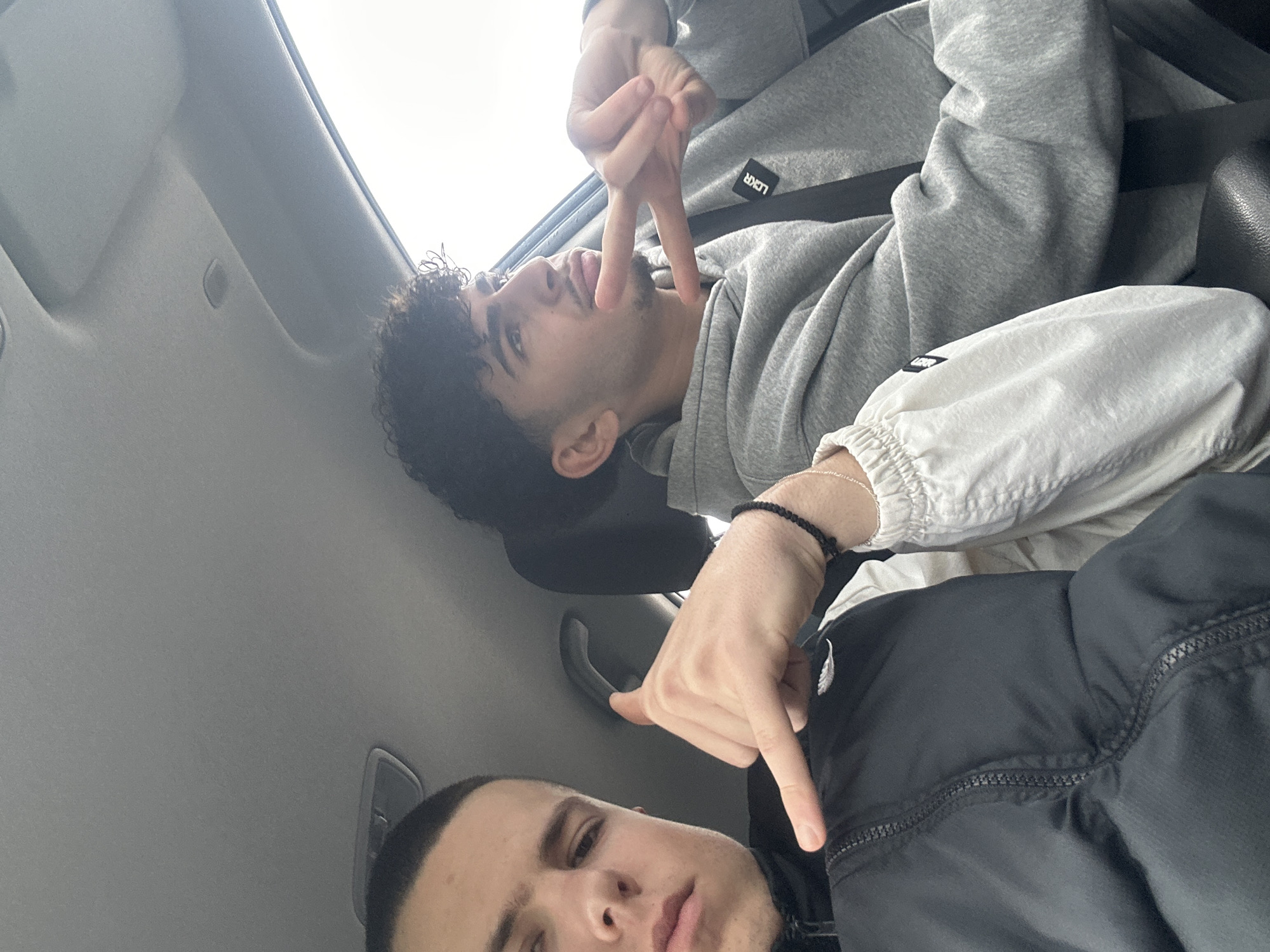 Two young men are in a vehicle. The interior suggests a car, specifically the roof and grab handle. One man, with dark curly hair, is wearing a light grey hoodie. The other man has short hair and is wearing a dark jacket. One man is making a hand gesture above the other's head. The location is unspecified, though the request mentioned Piacenza, Italy, there is no explicit confirmation of this.