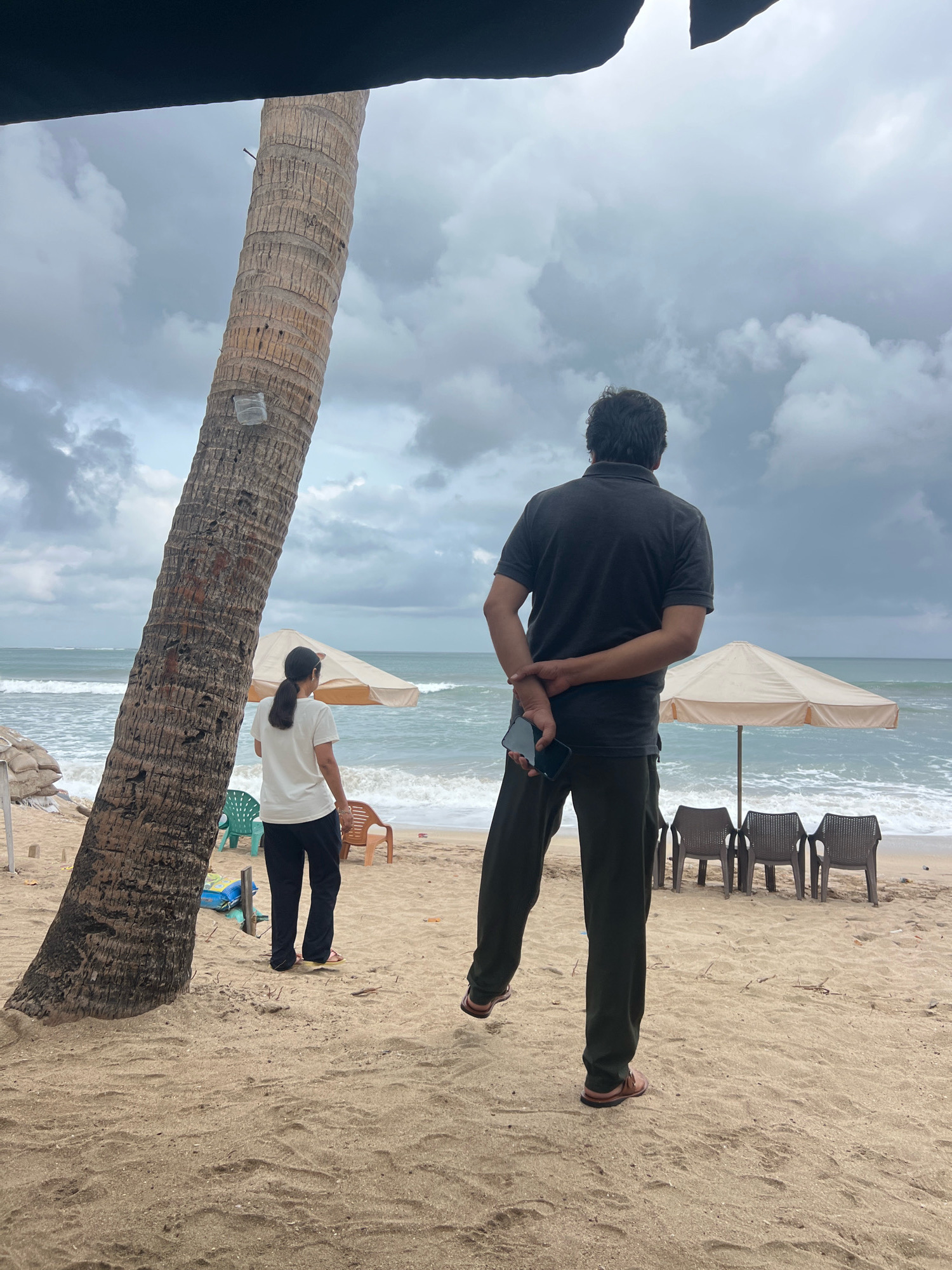 The image depicts a beach scene in Kuta, Indonesia, under a cloudy sky. In the foreground, a large coconut tree trunk stands to the left. To the right of the tree, a man is standing with his back to the camera, wearing a dark grey polo shirt and dark trousers. He is holding a black smartphone in his right hand, which is clasped behind his back. A woman in a white t-shirt and black pants stands further back, also facing away from the camera, to the left of the man.
The beach is sandy, with some dried leaves and twigs scattered on the surface. In the middle ground, there are several plastic beach chairs arranged in a row. Two beach umbrellas are set up behind the chairs, one partially visible to the left and a larger one in the center. Beyond the umbrellas, the ocean waves are breaking on the shore. In the far left background, there are some large sacks, possibly filled with sand or other materials, and a brightly colored plastic chair. The overall atmosphere is subdued due to the overcast weather.