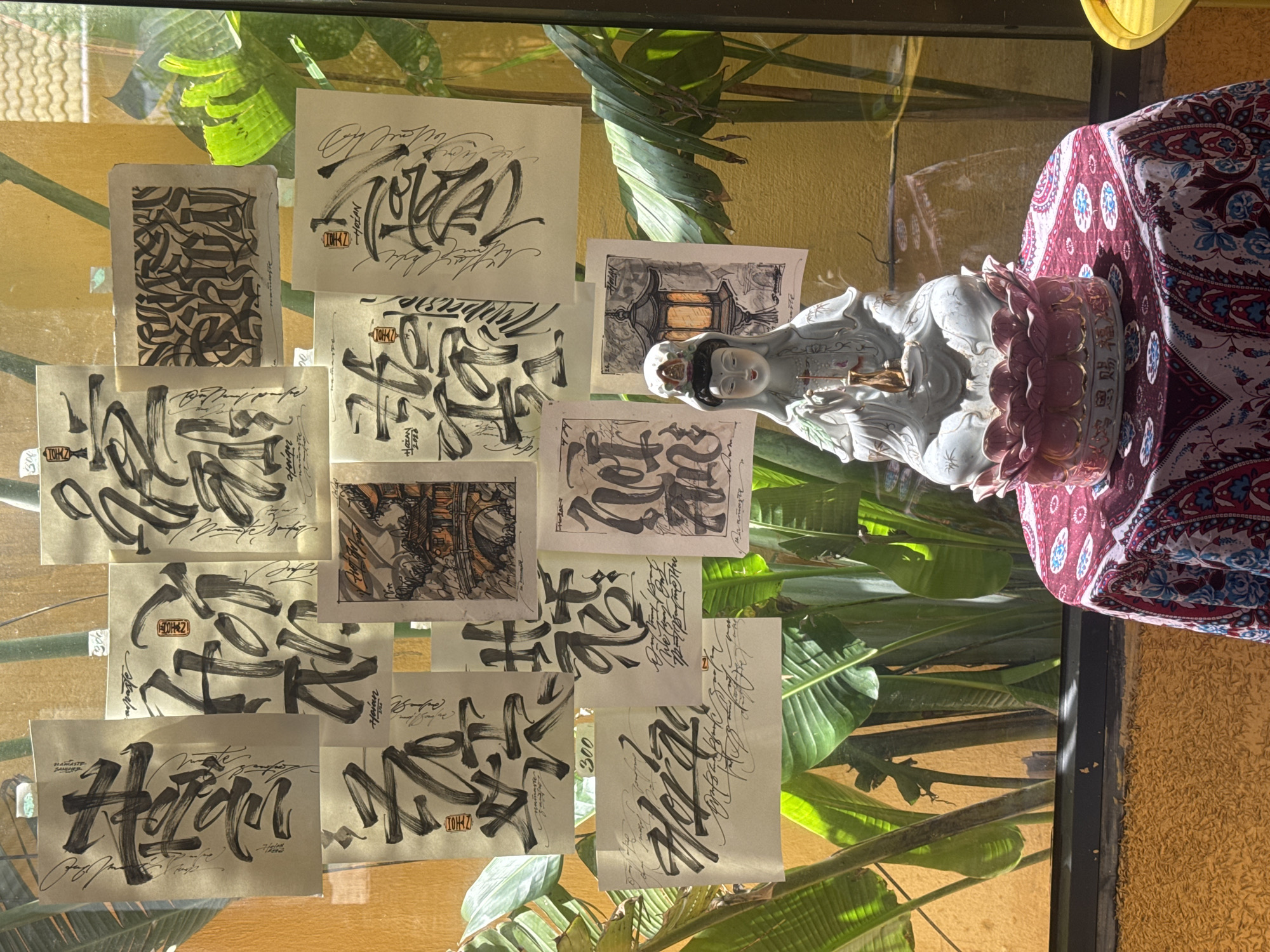 A collection of artwork, predominantly calligraphy and illustration on paper, is adhered to a glass surface. Visible among the art is a porcelain statue of a woman, dressed in pale blue robes, placed on a floral base, situated on a table draped with a patterned red and blue tablecloth. The setting appears to be an interior space with a window overlooking lush greenery. The overall impression is one of artistic display and cultural elements. Given the reference to "Namaste" and potentially Vietnamese calligraphy, the location may plausibly be in Đà Nẵng, Vietnam, although this is not directly indicated in the image.