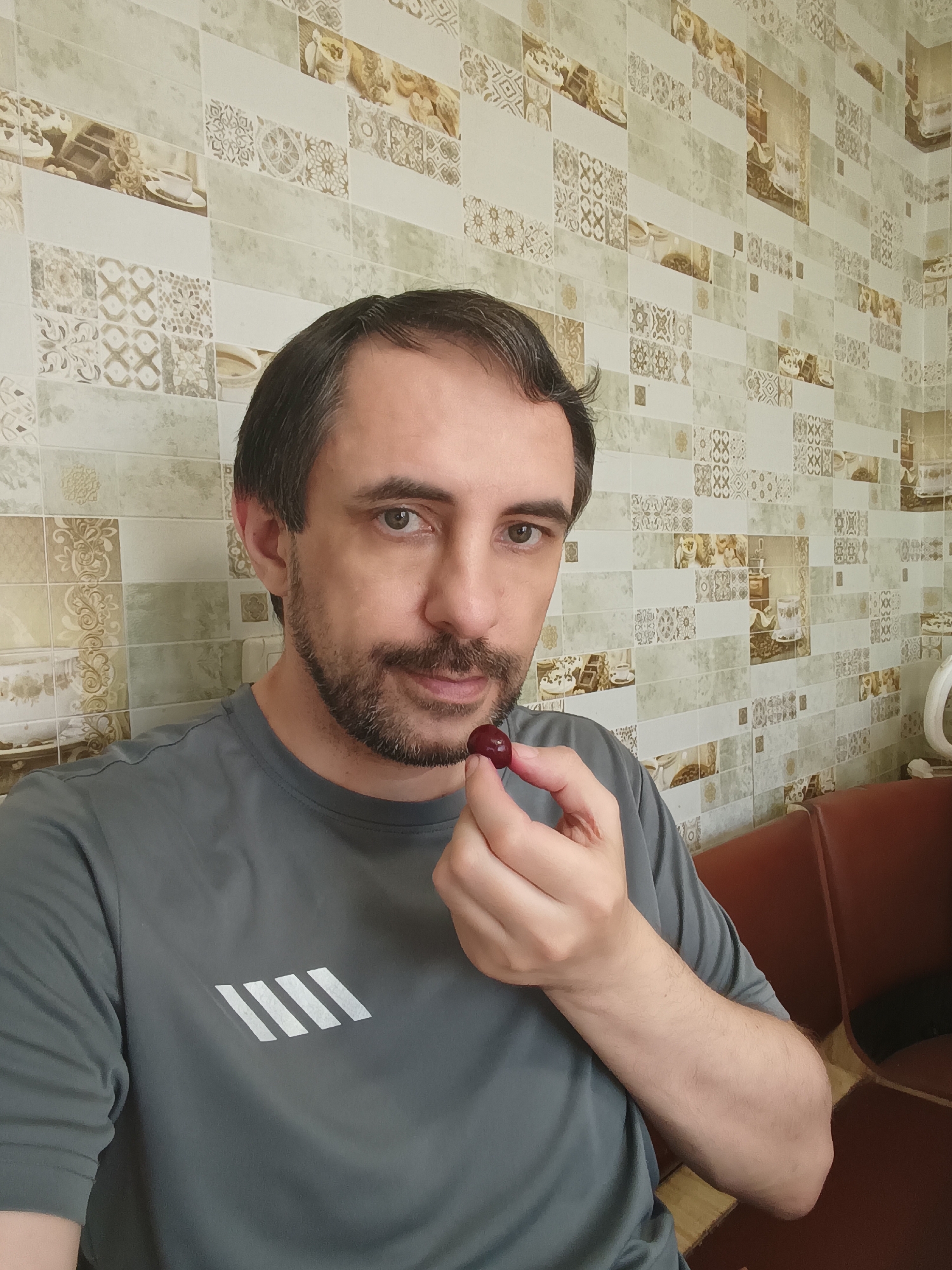 In this close-up selfie taken in Izmail, Ukraine, a man with short dark hair and a beard holds a single cherry between his fingers, bringing it towards his mouth. He is wearing a grey t-shirt with a white diagonal stripe logo on the left chest. The background features a tiled wall with a pattern of beige and off-white squares, some of which depict floral designs and images of coffee cups and baked goods. To the right of the man, a portion of a brown leather couch and a white kettle are visible. The lighting suggests an indoor setting, likely a kitchen or dining area.
