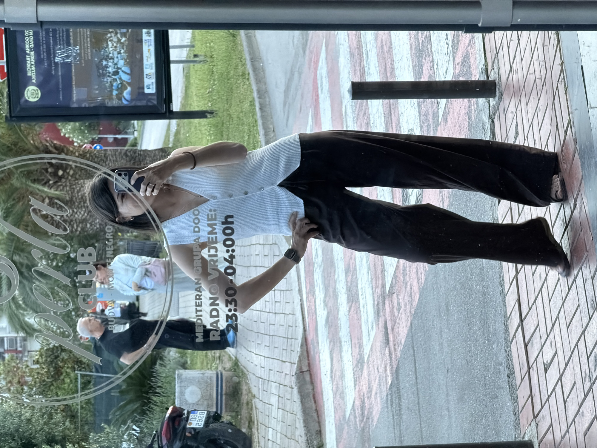 The image is a reflection, presumably in a glass door or window, showing a woman taking a picture of herself with a smartphone. She is wearing a light-colored top and dark pants.
The reflection also shows a partial view of the interior behind her, which appears to be the "Top Hill Club" in Budva, Montenegro. This is indicated by the lettering "Top Hill CLUB" and "BUDVA - MONTENEGRO" visible in the reflection. "MEDITERAN GRUPA DOO" and "RADNO VRIJEME: 23:30-04:00h" are also visible, indicating the business's opening hours from 11:30 PM to 4:00 AM.
Other people are also reflected in the image, along with some greenery and a motorbike parked near the establishment. The surrounding environment appears to be a street or pedestrian area with pavement markings.