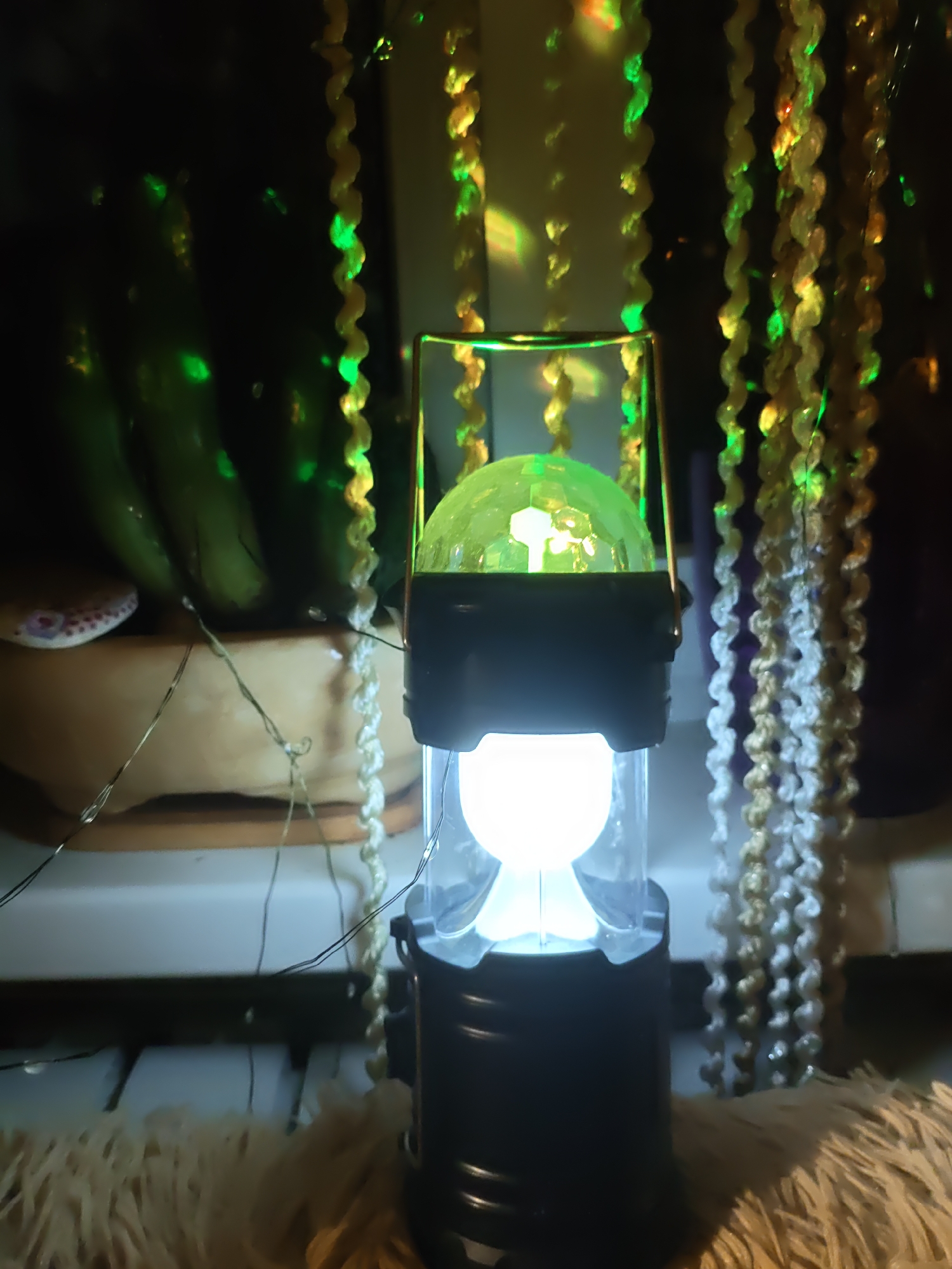 The image shows a portable LED lantern with a disco ball on top, emitting a green light. The lantern is positioned on a white surface, possibly a shelf or windowsill, in what appears to be an indoor setting. Behind the lantern, a dark background is illuminated by green lights, creating a festive or decorative atmosphere. There are also strands of decorative beads hanging vertically, some of which are also lit with green and yellow lights. In the lower foreground, a fluffy, tan-colored rug or textile is visible. To the left of the lantern, partially obscured by it, is a light-colored, curved object, possibly a bowl, with a decorative pattern on its surface. Green lights are also reflected on what appear to be several large, dark green objects in the background, which could be fruits or vegetables, such as cucumbers or gourds. The lighting suggests it is nighttime or the room is dimly lit. There is no indication of the city of Rostov-on-Don, Russia, or any specific location. There are no people visible in the image.