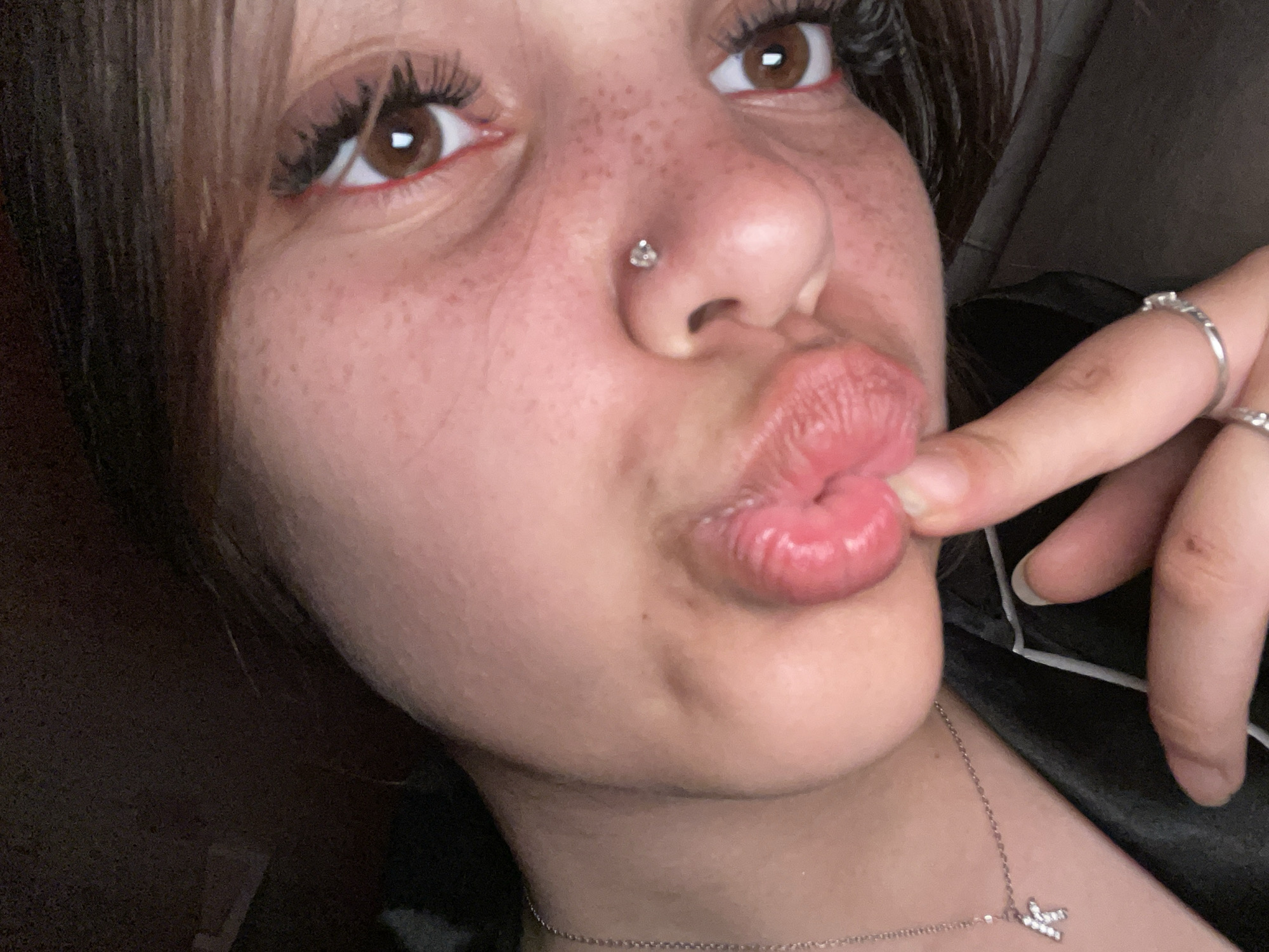 A close-up, low-angle portrait shows the face of a young person wearing makeup. The subject is making a pouty face, with their lips pursed and a finger touching them. A nose piercing with a small, jeweled stud is visible on the right side of their nose. Their eyes are a rich brown and are accentuated with dark eyeliner and mascara. Freckles are scattered across their cheeks and nose. A silver necklace with a "K" pendant is visible on their neck. The background is dark and out of focus, suggesting an indoor setting.
