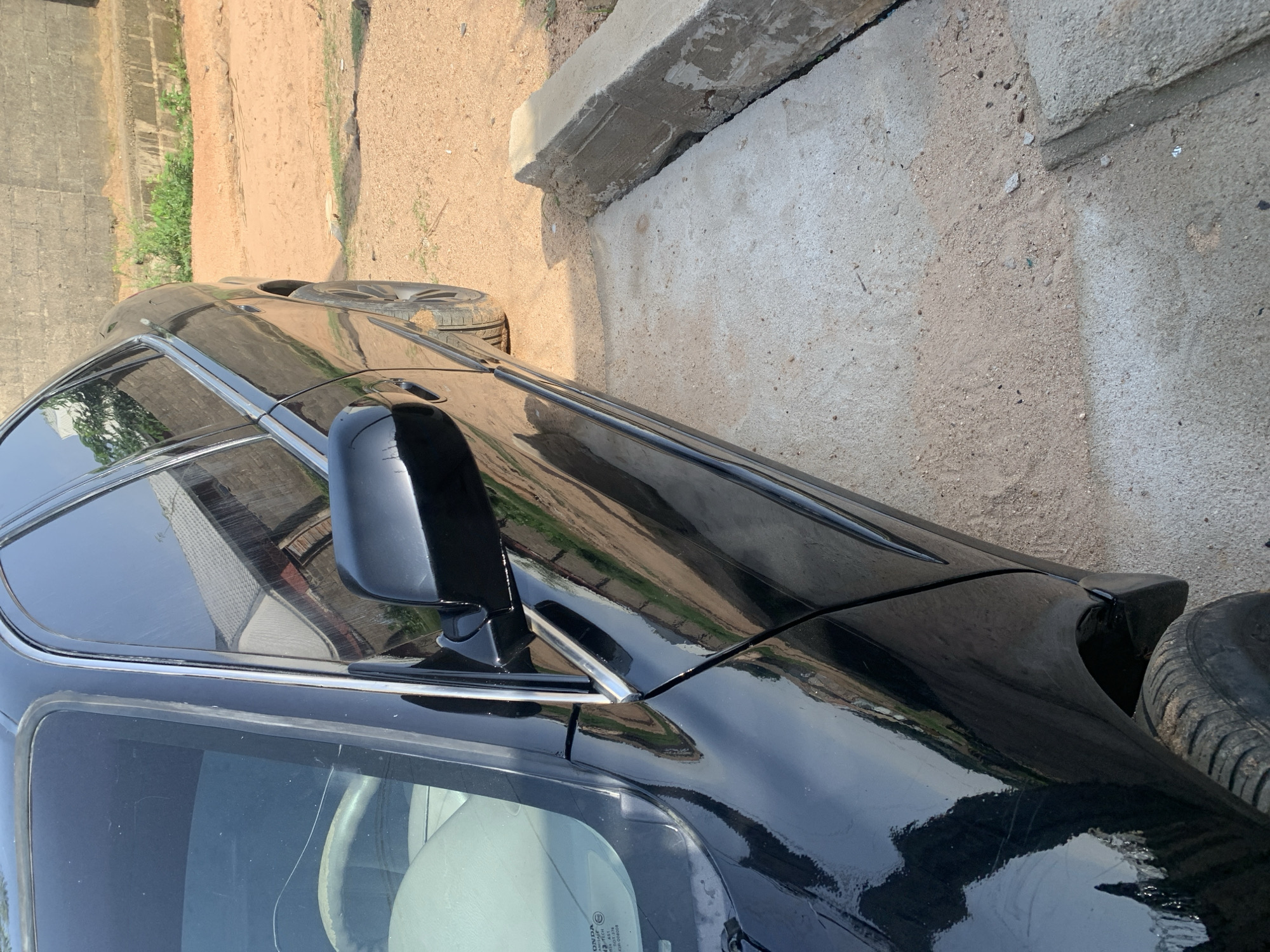 A black sedan is parked outdoors on a dirt and concrete surface. The vehicle is oriented facing left, with its right side visible to the camera. The car's shiny black exterior reflects the surrounding environment, which appears to be an urban or semi-urban area with buildings and vegetation. The ground is a mix of loose sand and a rough concrete slab, with a concrete barrier or curb present on the right side of the image. The lighting suggests it is daytime, with clear shadows cast by the car and surrounding objects. The scene appears to be static, with no human activity depicted.