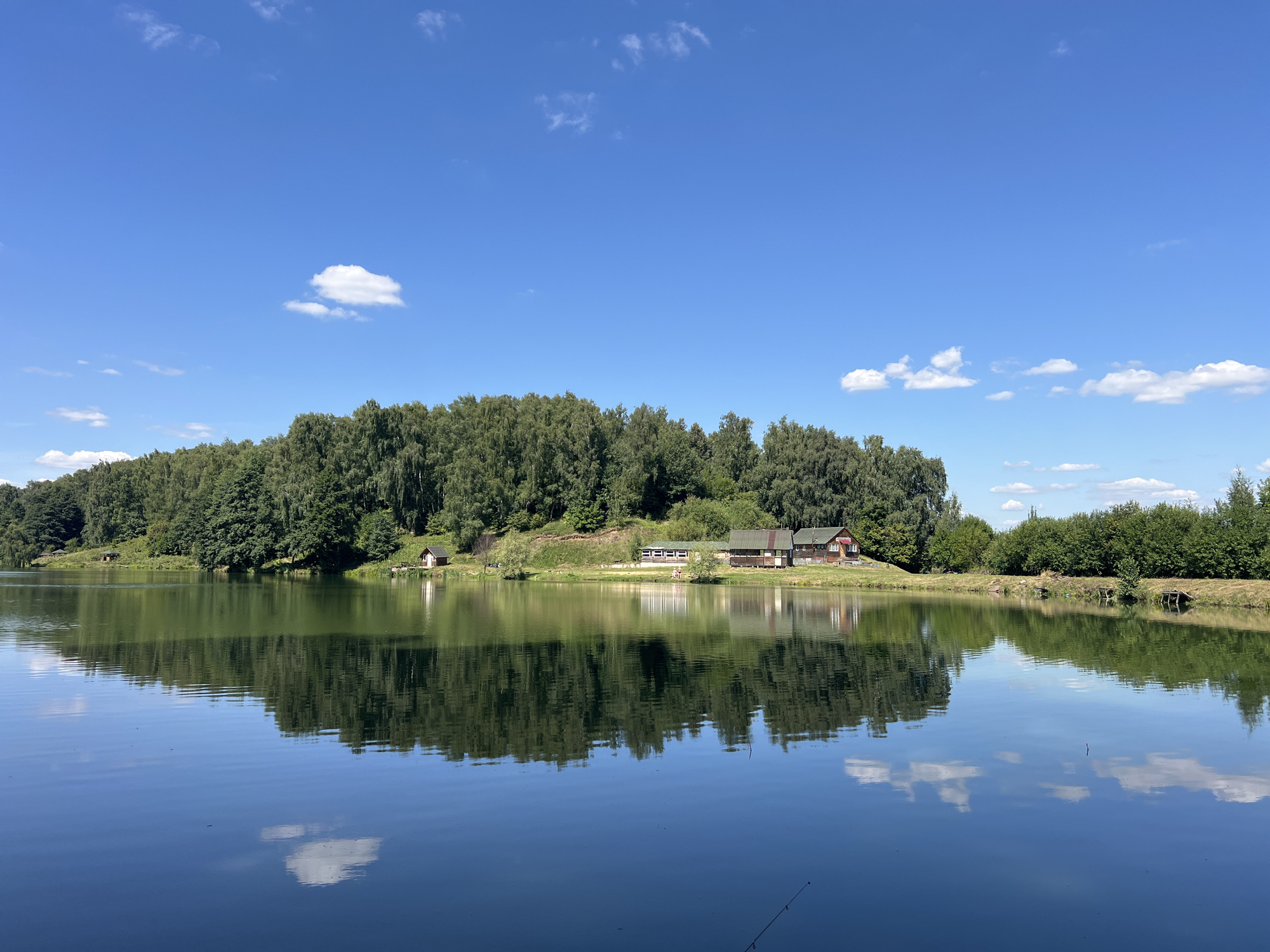 This image captures a serene landscape scene at the Мосметаллоконструкция Онт, Russia. A calm body of water dominates the foreground, its surface reflecting the clear blue sky dotted with white clouds, and the lush green treeline on the opposite bank. Along the water's edge, nestled amongst the trees, are several buildings. The main structure appears to be a lodge or a series of cabins, with a red roof and wooden siding, situated on a gentle slope. Smaller structures, possibly sheds or outbuildings, are also visible. The trees are dense and varied, creating a thick, verdant backdrop to the water. The overall impression is one of tranquility and natural beauty, likely a recreational or residential area by the lake.