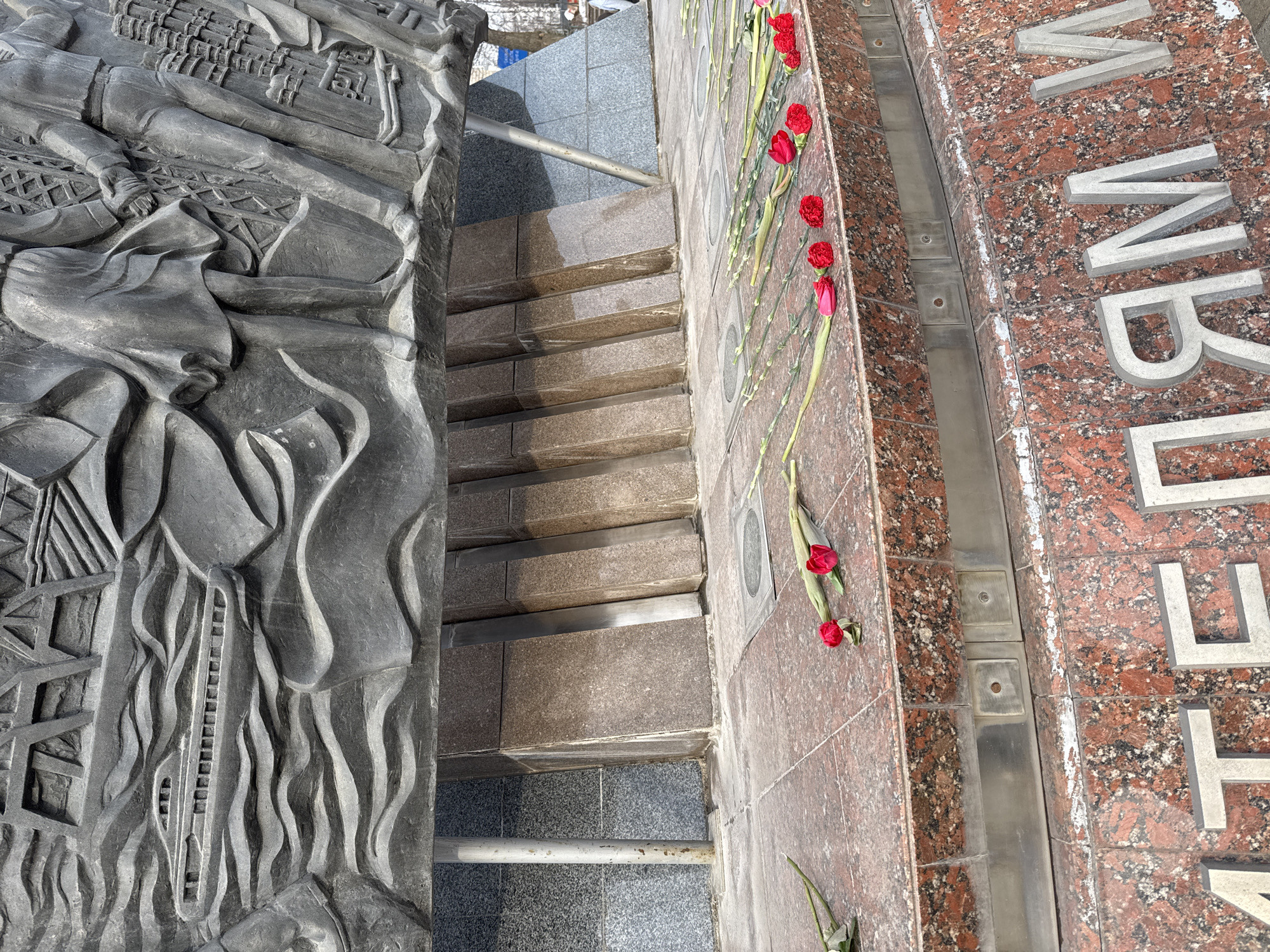 This image shows a stone staircase with a metallic handrail, placed between a stone wall with carved figures on the left and a red granite wall with white letters on the right. Red tulips are laid on the granite wall. The location is Unknown, Unknown.
