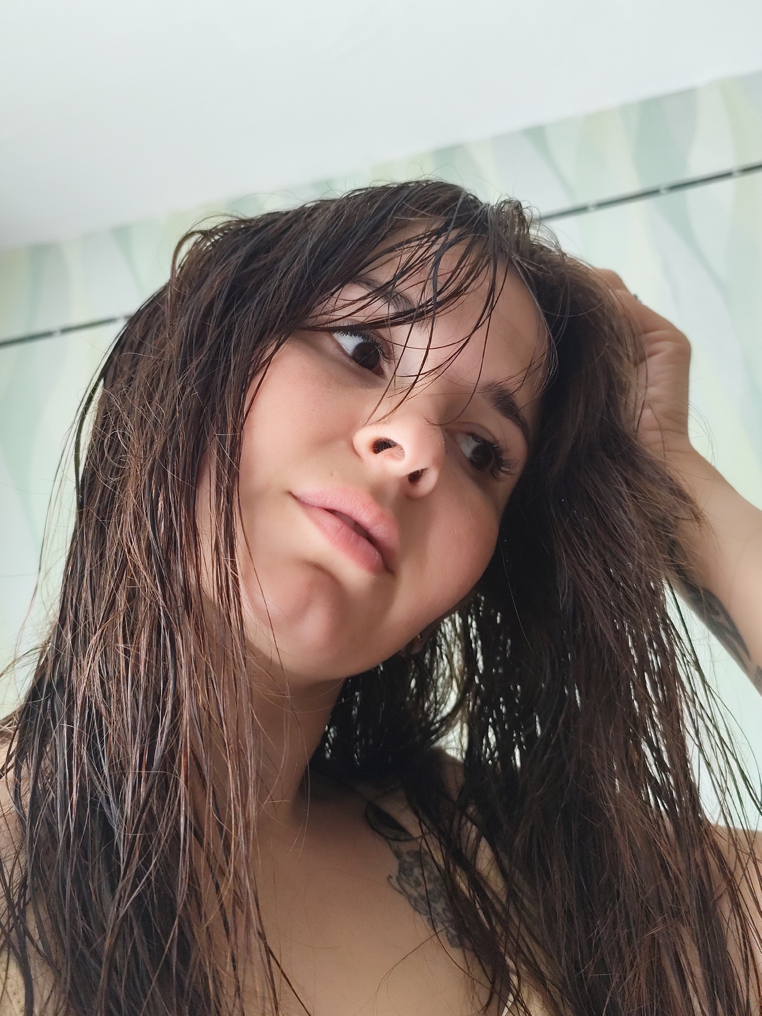 A young woman with long, wet dark brown hair is looking slightly downwards and to the left of the frame. She is touching her hair with her right hand. The woman appears to be indoors. The background includes a light-colored wall with a subtle pattern. There is no information available to determine if this is in the city of Фоки, Russia.
