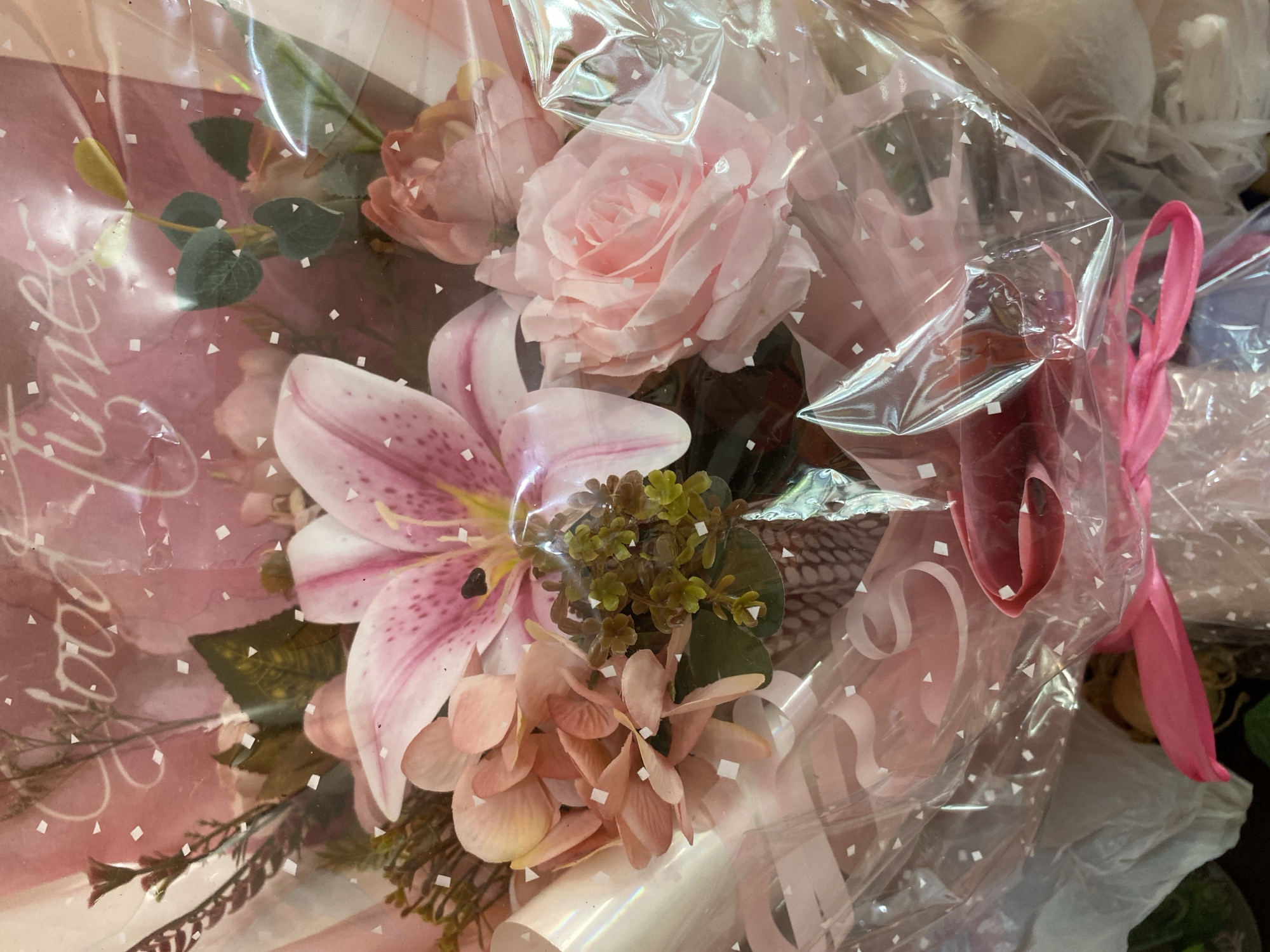 This is a close-up, eye-level shot of a bouquet of pink and white flowers wrapped in clear plastic. The bouquet is composed of a large pink lily with dark spots, several pink roses, and other smaller pink and green floral elements. The plastic wrapping has a pattern of small white squares and triangles. The background is a pink paper with the handwritten text "Good Times" in white cursive script, also with the small white square and triangle pattern. A bright pink ribbon is tied around the bottom of the bouquet. The image does not provide any information about the location in Lambaro Kueh, Indonesia.