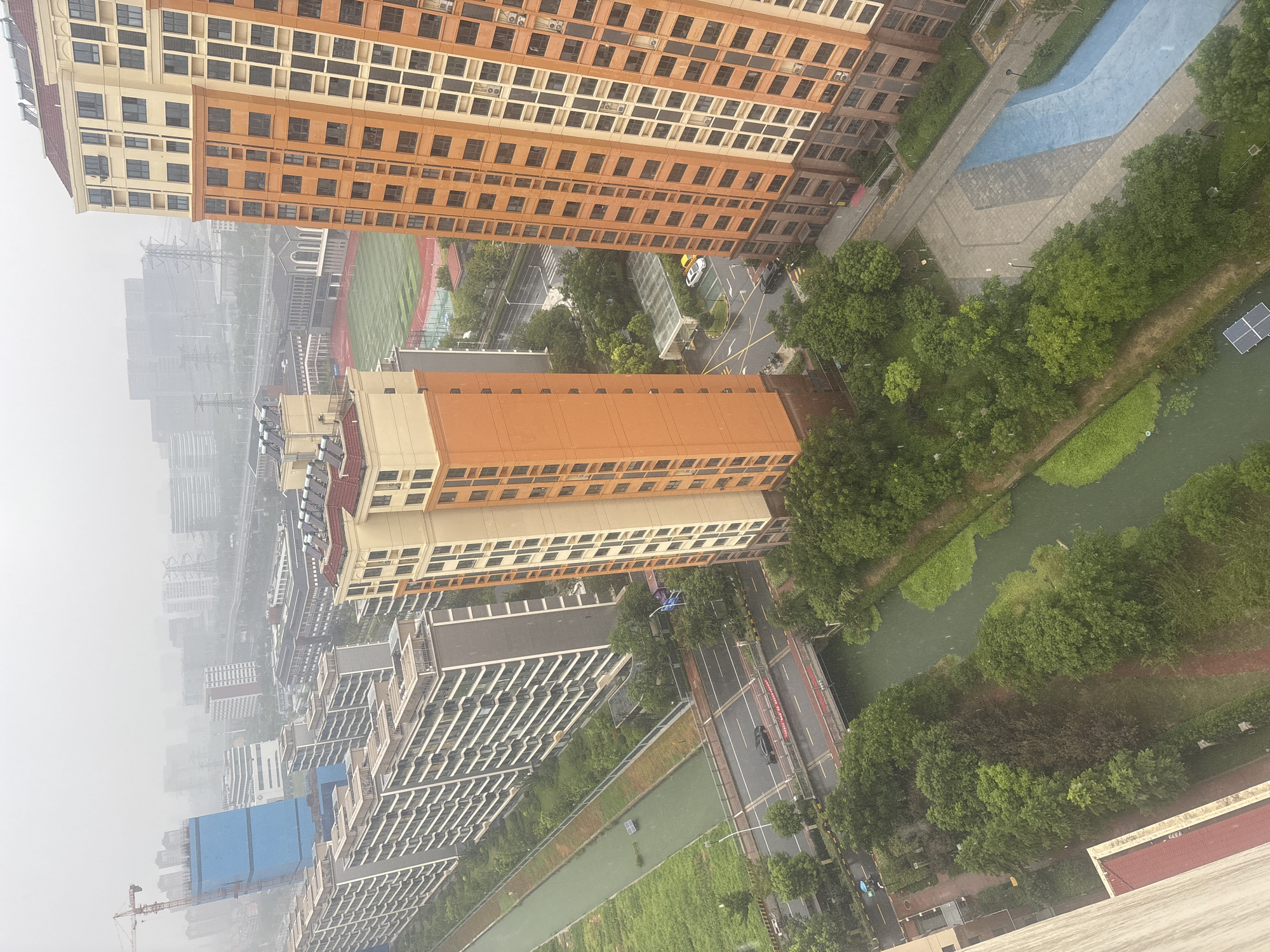 This aerial, high-angle image captures a hazy, overcast day in Suzhou Industrial Park, China. The scene is dominated by several large, multi-story residential buildings, two of which are prominently featured in the foreground. The building on the right is a tall, orange and cream-colored apartment complex with numerous windows. To its left, a slightly shorter, orange and beige building stands, also featuring many windows and a red-tiled roof.
Between these buildings and further into the distance are rows of other apartment buildings, appearing mostly gray and white, with balconies. A blue-clad construction site with a crane is visible in the upper left quadrant. A green, rectangular sports field with red running tracks surrounds it.
A body of water, appearing murky green, runs horizontally through the mid-ground, bordered by lush green trees and vegetation. A road with traffic crosses this waterway via a red bridge in the middle left of the image. Cars are visible on the road and a solar panel is floating in the canal. A paved walkway or plaza area with a blue design element is situated to the right of the orange buildings in the foreground. The overall impression is of a densely populated urban residential area with accompanying infrastructure.