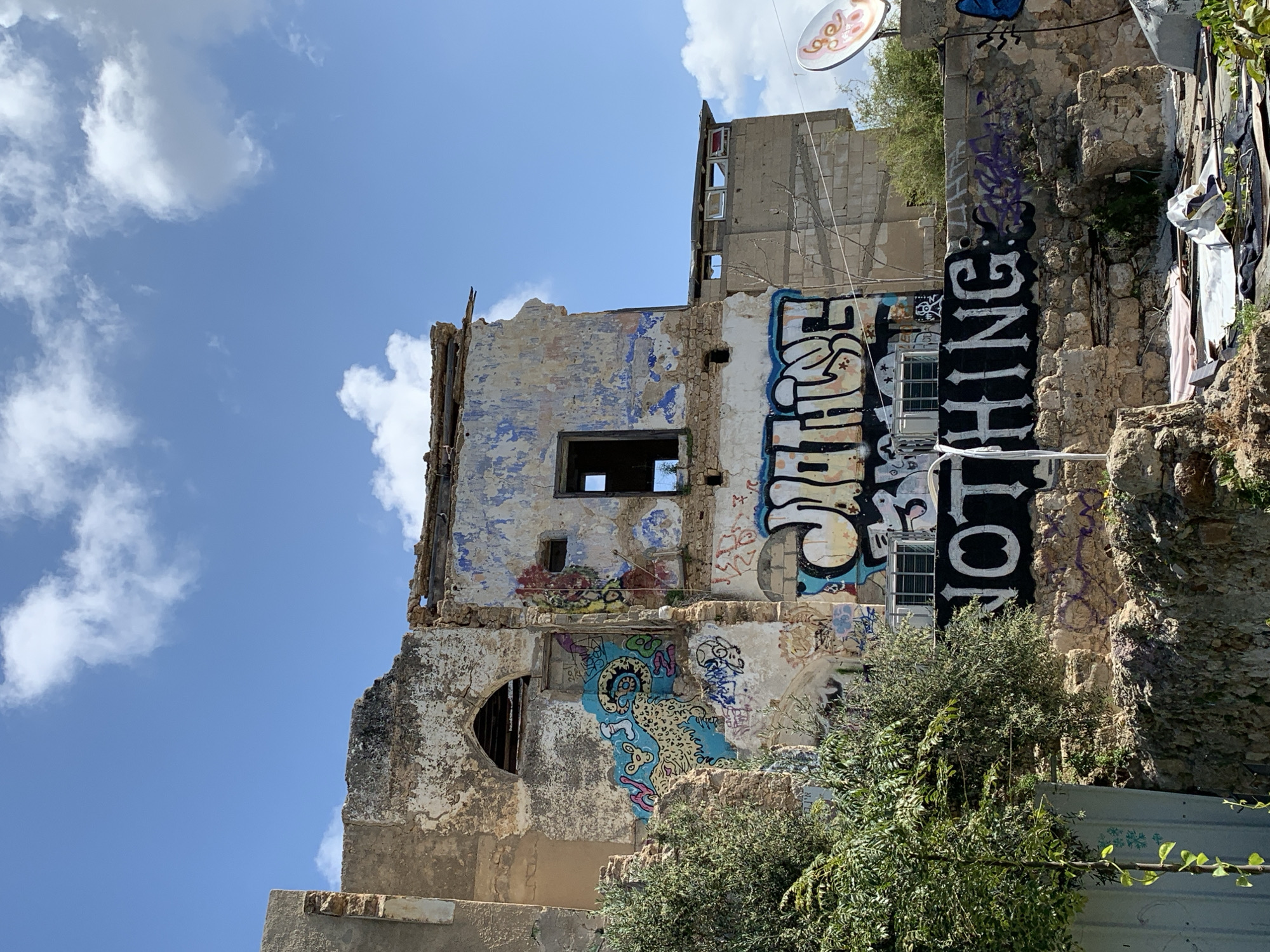 The image shows the remnants of a crumbling building covered in graffiti in Tel Aviv, Israel. The facade is largely deteriorated, with exposed brick and crumbling plaster. Graffiti covers much of the wall surface, with one section displaying the word "NOTHING" vertically. The sky is partly cloudy. Vegetation obscures some parts of the lower facade.
