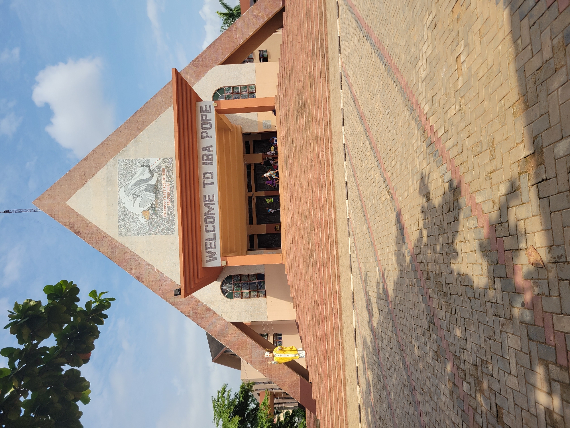A full shot shows the front of a church with a large, peaked roof. The roof is adorned with a mosaic of a kneeling figure and text that reads "I AM HAPPY TO SET FOOT ON YOUR BELOVED LAND." Below the mosaic, a sign reads "WELCOME TO IBA POPE." A wide set of stairs leads up to the entrance. A statue of a figure in a yellow robe and white mitre stands on the left side of the stairs. Several people are gathered near the entrance. The foreground is a paved area with a pattern of interlocking bricks. Trees and a blue sky with clouds are visible in the background.