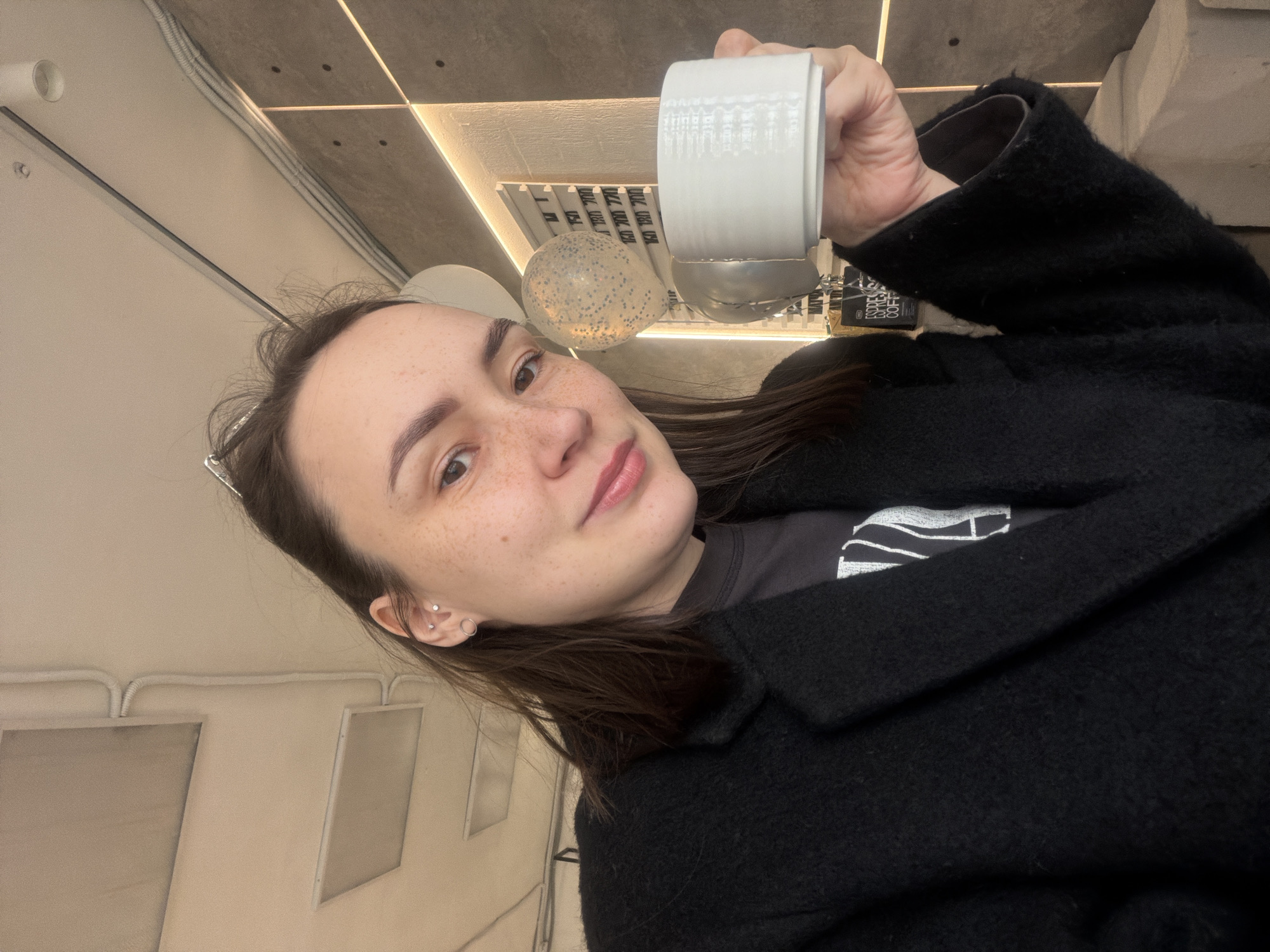 This tilted selfie captures a young woman inside what appears to be a modern cafe or retail space in Chaikovsky, Russia. The woman, with long dark hair, freckles across her nose and cheeks, and light brown eyes, looks directly at the camera with a subtle, pleasant expression. She has two silver stud piercings and a silver hoop earring on her visible left ear.
She is wearing a dark, textured coat over a dark t-shirt. In her right hand, she holds a white ceramic mug with vertical ridges, partially filled with a dark liquid, raising it towards the camera.
The background reveals contemporary interior design elements, including a concrete ceiling with integrated linear LED light strips and other light fixtures. White rectangular panels are mounted on a wall to her left. A translucent balloon filled with dark speckles is visible in the middle ground. Further in the background, a display stand can be seen with products, one clearly labeled "ESPRESSO COFFEE" on dark packaging. The overall setting suggests an urban, chic environment.