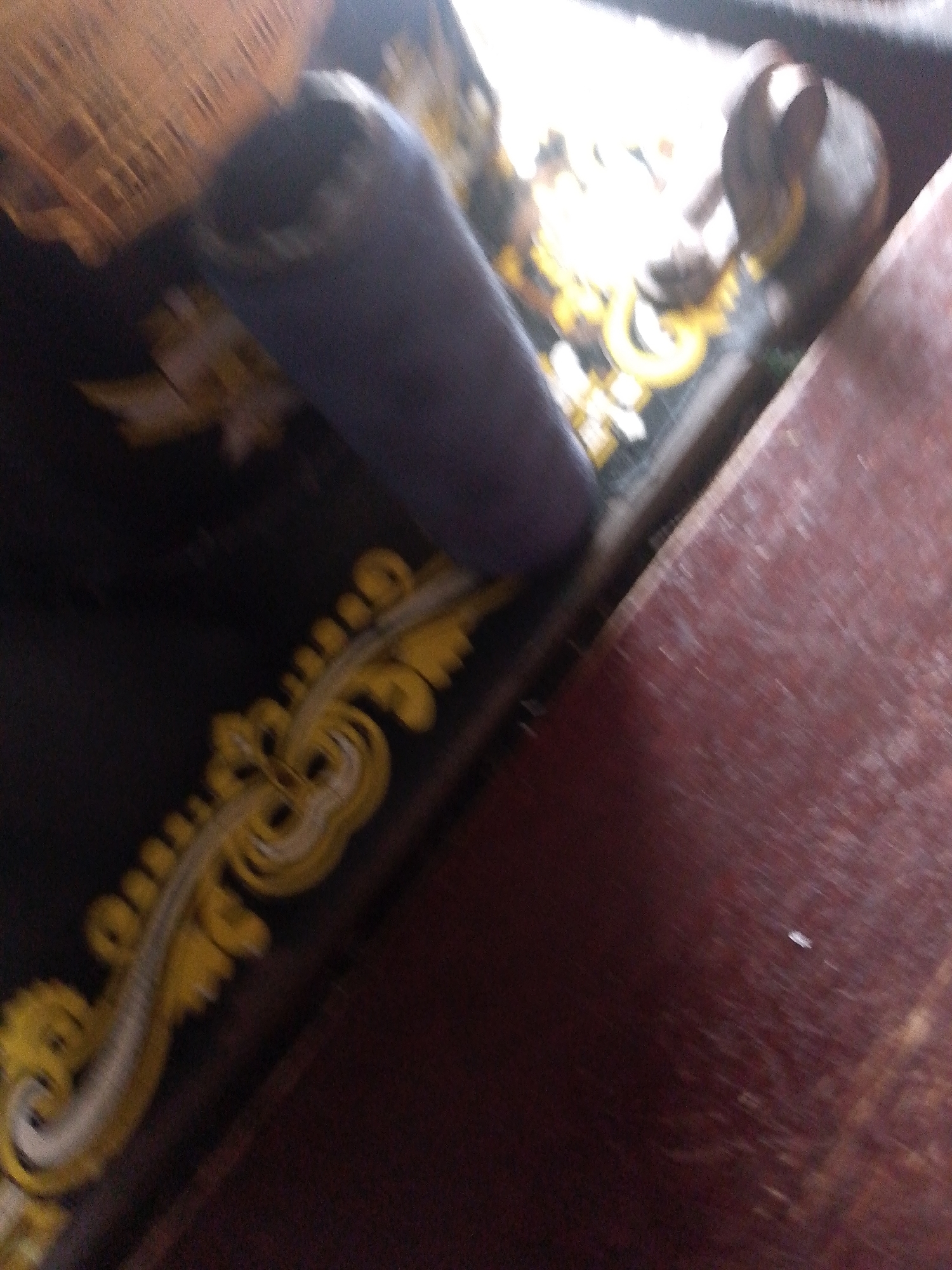 The image shows a close-up, angled view of a sewing machine. The machine's base is decorated with an ornate yellow and white scrollwork pattern. A dark blue fabric sleeve or cover is partially obscuring the machine. The background is dark and out of focus, with a reddish-brown surface to the right. The scene appears to be indoors. There are no people or specific actions visible. The location within Jauro Yinu, Nigeria, is not discernible from the image.