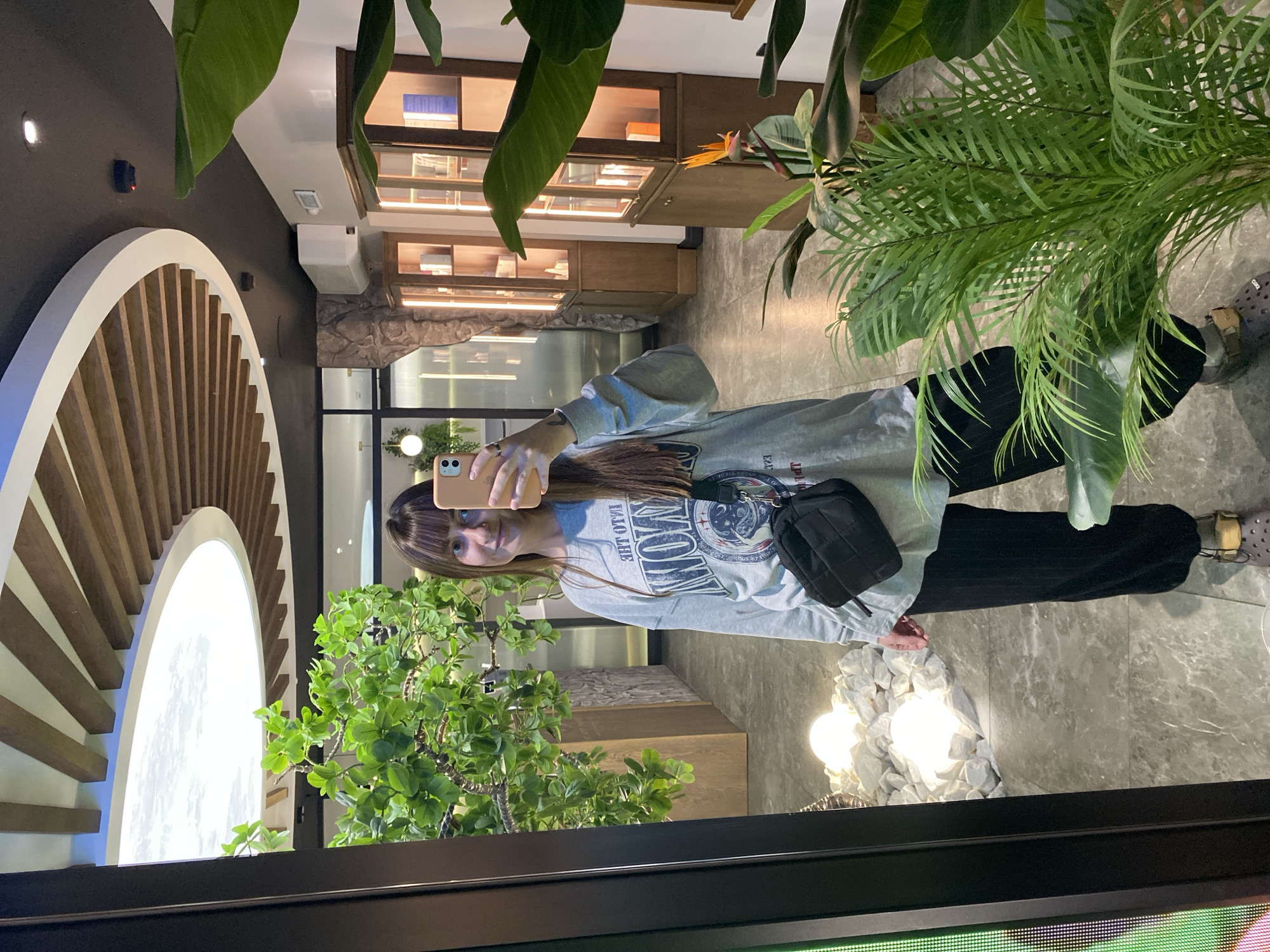 A woman with brown hair and bangs is taking a selfie with a tan-colored phone. She is wearing a gray sweatshirt with the word "VOTKINSK" written backward on it, black pants, and slip-on shoes. A black crossbody bag is slung over her shoulder. She is standing on a gray marble floor in what appears to be a building in Votkinsk, Russia. There are green plants surrounding her, including one that is close to the camera. In the background, there are display cases, a decorative arch, and light fixtures resembling rocks.