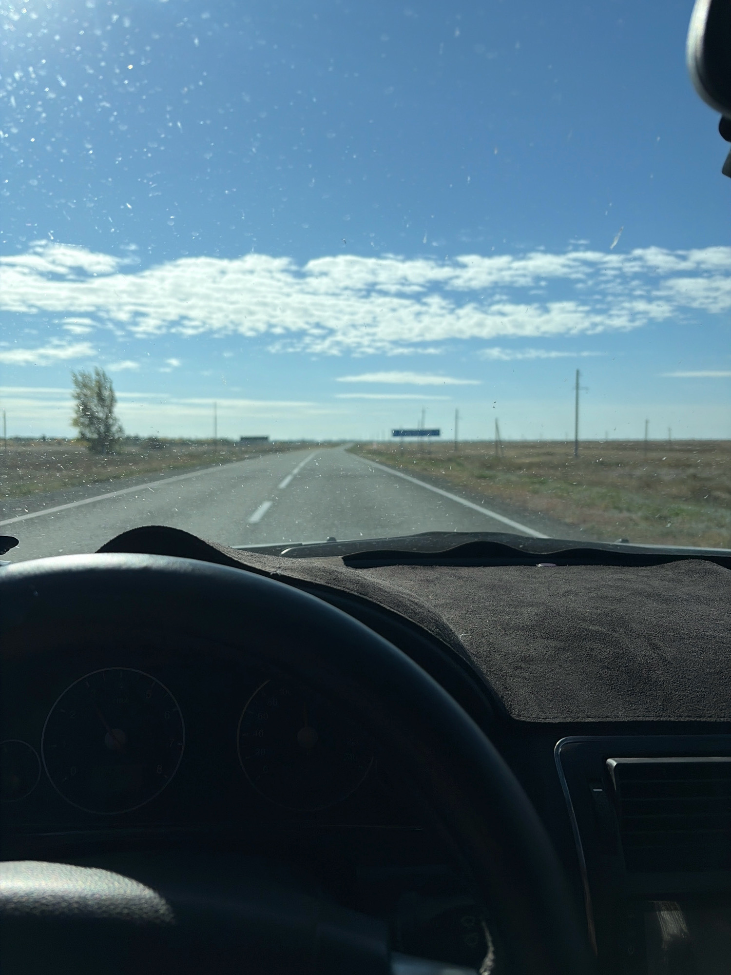 The image is taken from inside a vehicle, looking out through the windshield. The windshield is speckled with dust or water droplets. A road stretches into the distance, cutting through a flat, grassy landscape. The sky is a bright blue with scattered white, wispy clouds. Several utility poles are visible along the side of the road, and in the far distance, there appears to be a rectangular blue sign. A lone, windswept tree stands to the left of the road, further in the background. The dashboard and steering wheel of the vehicle are visible in the foreground. The dashboard has a dark, textured cover. Partially visible on the dashboard are two circular gauges, likely part of the car's instrumentation. The overall impression is of a drive through a rural or steppe-like environment under a clear sky. Given the context provided, the scene is likely located in or near Көктүбек ауылдық округі, Kazakhstan.