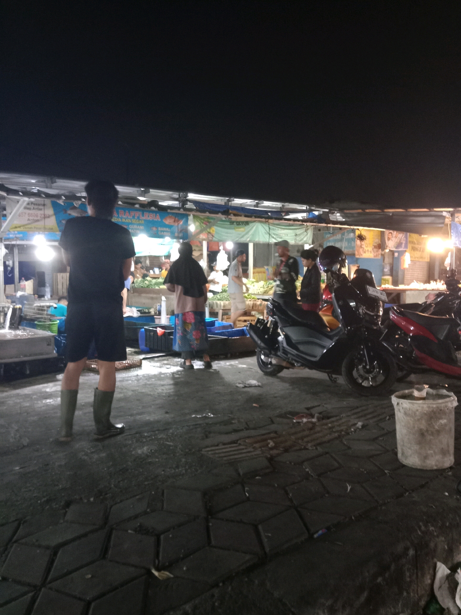 The image depicts an outdoor market scene at night in the city of South Tangerang, Indonesia. In the foreground, a person wearing a black t-shirt, black shorts, and green rubber boots stands facing away from the camera. To the right, a black scooter and a red motorcycle are parked. In the middle ground, several individuals are visible, including a person wearing a dark headscarf and a patterned skirt, standing near a stall. Another person in a black jacket and a person in a light-colored shirt are also present. The background shows market stalls with various goods, some of which appear to be vegetables. Overhead lighting illuminates the scene. The ground is paved with interlocking blocks and shows signs of debris.