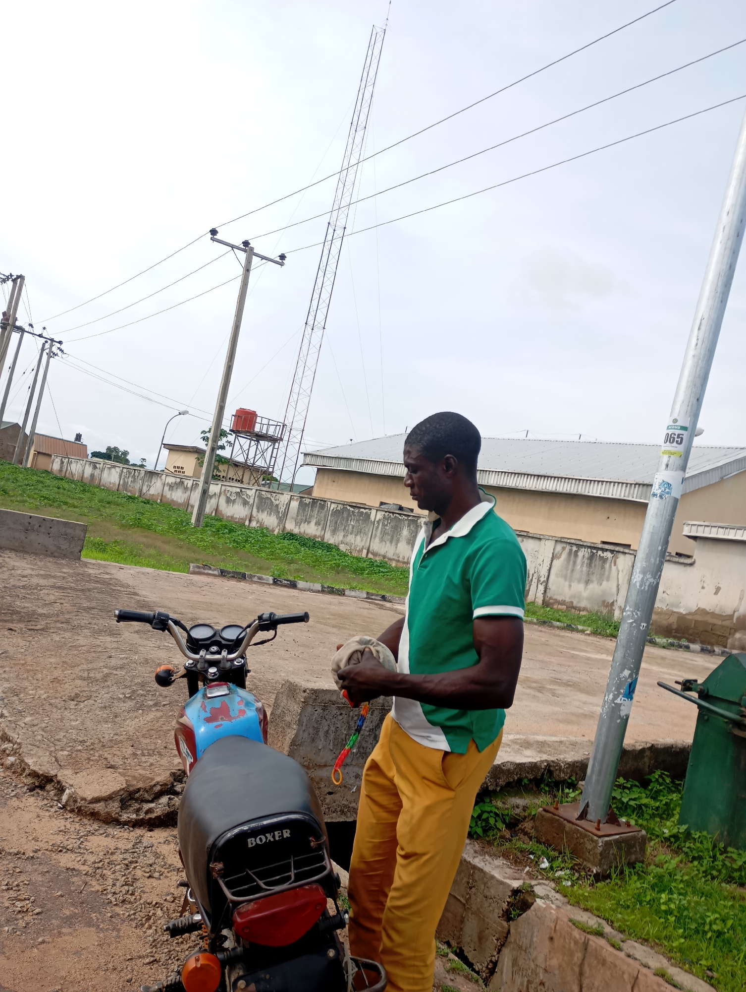 The image depicts an outdoor scene in Shendam, Nigeria. A man in a green and white polo shirt and yellow pants stands next to a motorcycle. He is holding a light-colored object in his hands, possibly a ball or a piece of cloth, with a colorful string attached to it. The motorcycle is parked on a dirt and concrete surface. In the background, there is a fence, a building with a red water tank on top, and a tall lattice tower structure. Power lines are visible overhead, supported by utility poles. The sky is overcast.