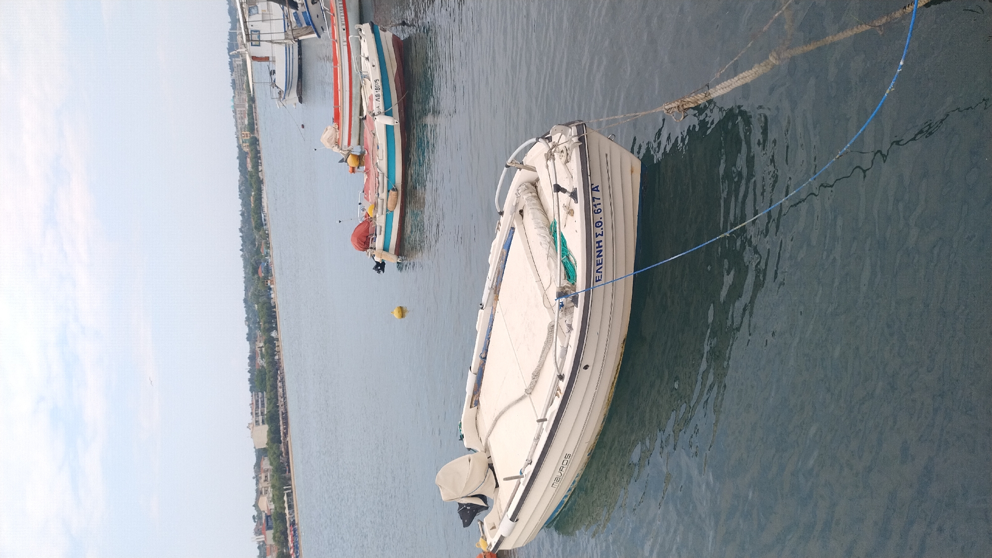 The image depicts a waterfront scene in Neoi Epivates, Greece. In the foreground, a white motorboat with a covered engine is moored in calm, dark blue water. The boat has blue trim along its hull and the registration "ΕΛΕΝΗ Σ.Ο. 617 Α'" is visible in blue lettering on the stern. Another white boat with the name "MAVROS" is visible on the side of the boat. Further back, another motorboat, predominantly white with blue and red accents and a red cover over its engine, is also moored. A yellow buoy floats nearby. In the background, the coastline of Neoi Epivates is visible, featuring a sandy beach lined with umbrellas and chairs, a strip of trees, and buildings. The sky is overcast with pale blue and white clouds. The overall scene is one of a quiet harbor or bay, with several boats at rest.