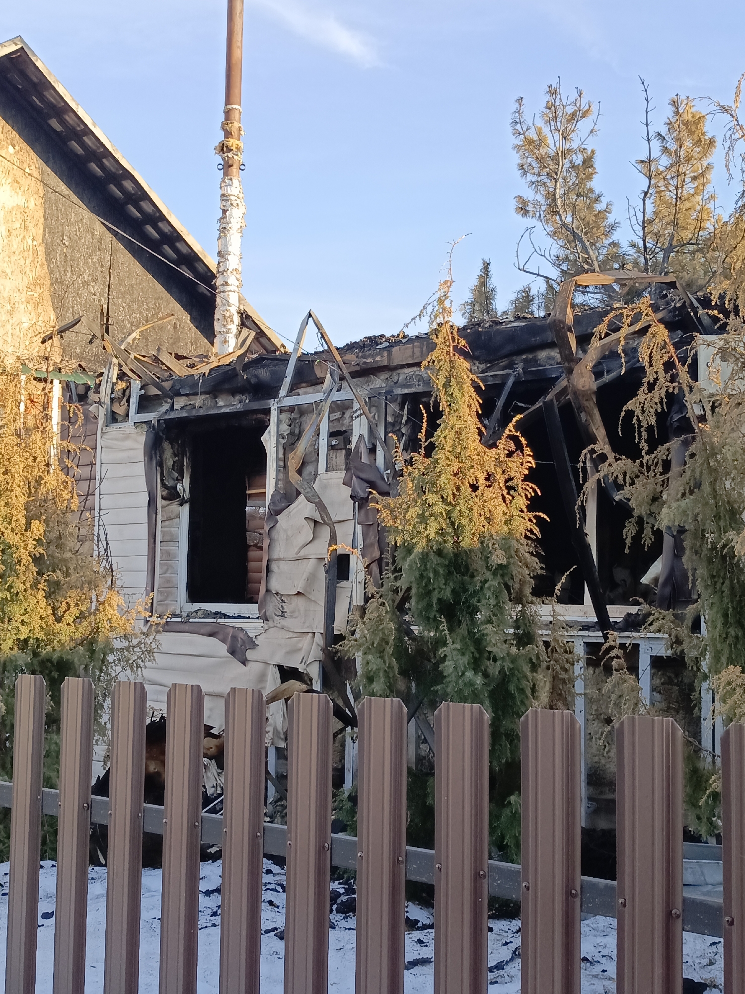 A severely fire-damaged residential structure stands behind a brown picket fence. The building's facade is partially collapsed, revealing charred beams and remnants of siding. A rusty chimney pipe extends from the compromised roof. Trees and shrubs partially obscure the view of the house. The ground is covered with what appears to be snow or ice. The sky is a clear blue. This is reportedly located in Chaikovsky, Russia.