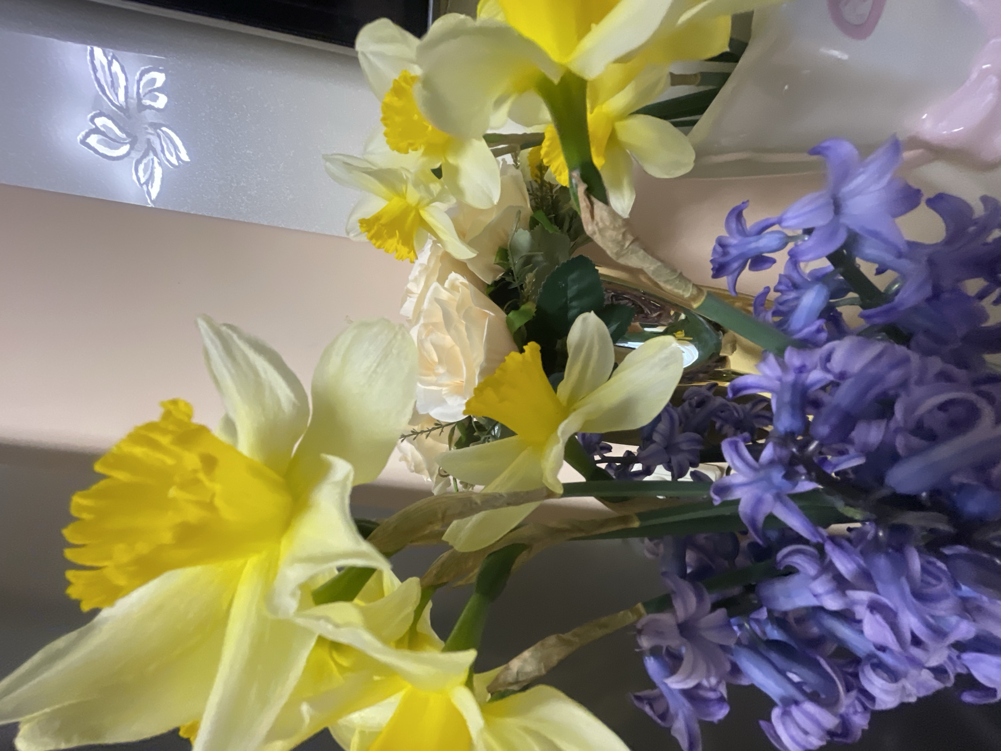 A close-up shot of a bouquet of flowers, including daffodils with yellow and white petals, white roses, and purple hyacinths. The background is a neutral beige color, with a decorative white object with a floral design visible on the left. The image appears to be taken indoors under artificial lighting. No people are visible. The location is not apparent from the image.