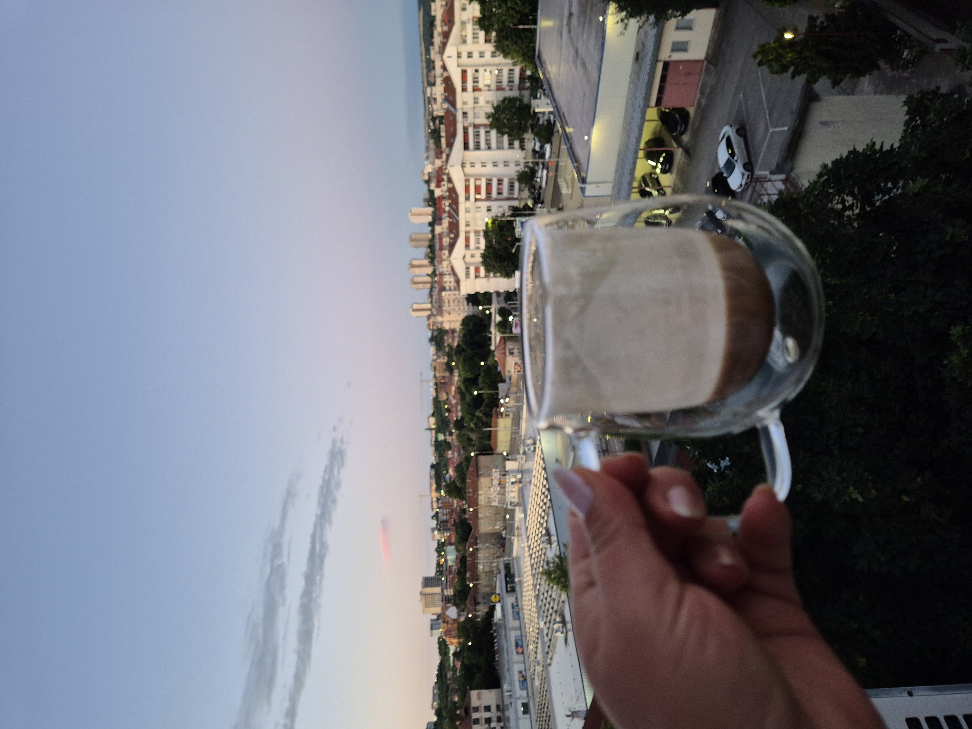 A close-up shot shows a hand holding a double-walled glass mug filled with a beverage, possibly coffee, against a blurred cityscape at dusk. The mug is positioned in the foreground, obscuring a portion of the view. In the background, buildings with red-tiled roofs are visible, along with taller apartment blocks and what appears to be a supermarket with a yellow "Lidl" logo. The sky is a soft gradient of blue and pink, indicating either sunrise or sunset. Several cars are parked in an outdoor lot in front of some buildings. The overall scene suggests a relaxed moment with a drink, observing the city from a higher vantage point.