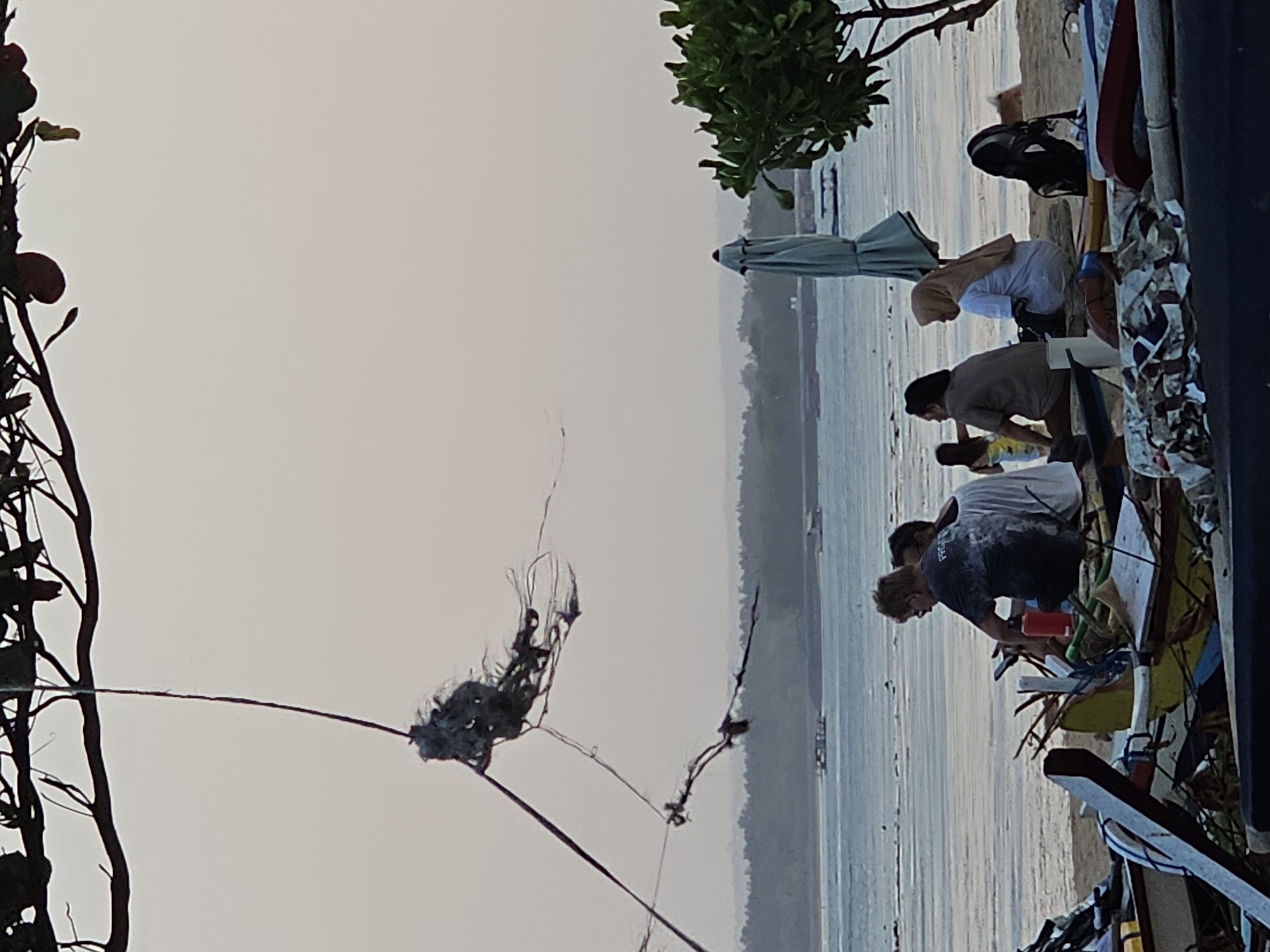 A group of at least six individuals are seated on a beach in Denpasar, Indonesia, near the shoreline. The time of day appears to be either dawn or dusk, given the soft, diffused light and the pale sky. In the foreground, several overturned and damaged small boats or fishing vessels are piled on the sand, some with debris scattered around them. A large, pale blue umbrella stands closed to the right of the group. Beyond the beach, the calm sea stretches towards a distant, hazy coastline lined with trees. In the distance, a few small boats are visible on the water. The overall atmosphere is serene and relaxed.