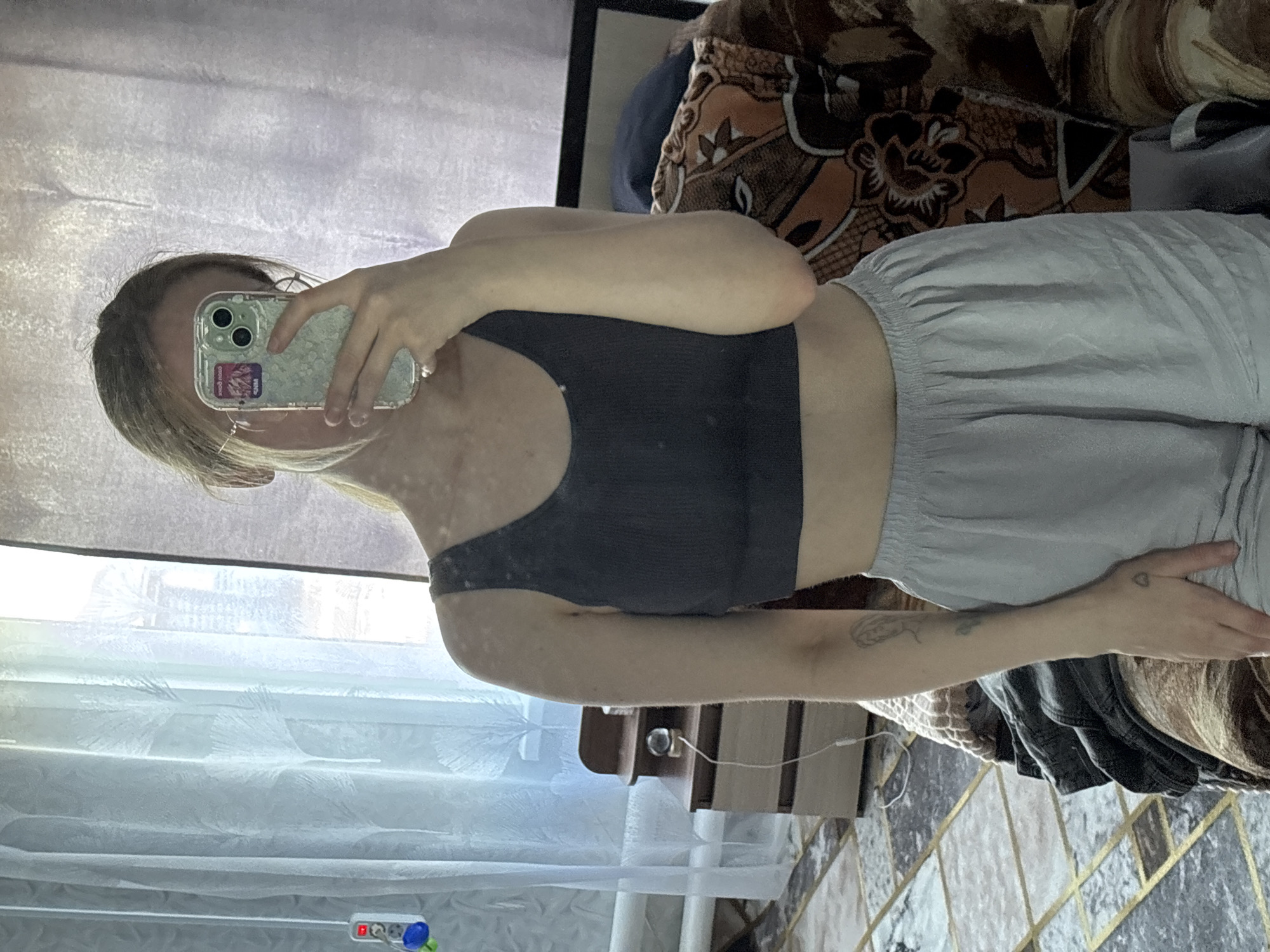 The image shows a young woman taking a selfie in a mirror. She is wearing a dark-colored sports bra and light gray shorts. Her blonde hair is pulled back. She is holding a smartphone in her right hand. The background appears to be a room with a patterned couch and curtains. The lighting suggests it might be daytime. There are no discernible emotions or specific activities taking place beyond the act of taking a photograph. The image is oriented vertically, with the subject's head towards the left and her legs towards the right.