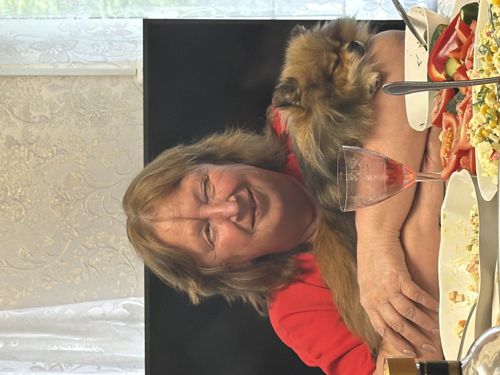 This media file features a woman with blonde hair and fair skin, wearing a red top, and a small fluffy dog resting on her shoulder. She appears to be smiling with her eyes closed, suggesting a moment of joy or relaxation. The setting seems to be indoors, possibly at a dining table, indicated by the presence of food dishes and a wine glass. The food dishes contain what looks like salad and vegetables, including tomatoes and bell peppers. A wine glass, partially filled with a pink liquid, is also visible. The background shows a window with white curtains and a patterned wallpaper. The overall mood is warm and convivial. The woman's relaxed posture and the presence of the dog suggest an informal gathering or a moment of leisure. The scene is likely taking place during the daytime, given the natural light coming from the window. There is no visible text in the image. The location context is Бронница, Russia.