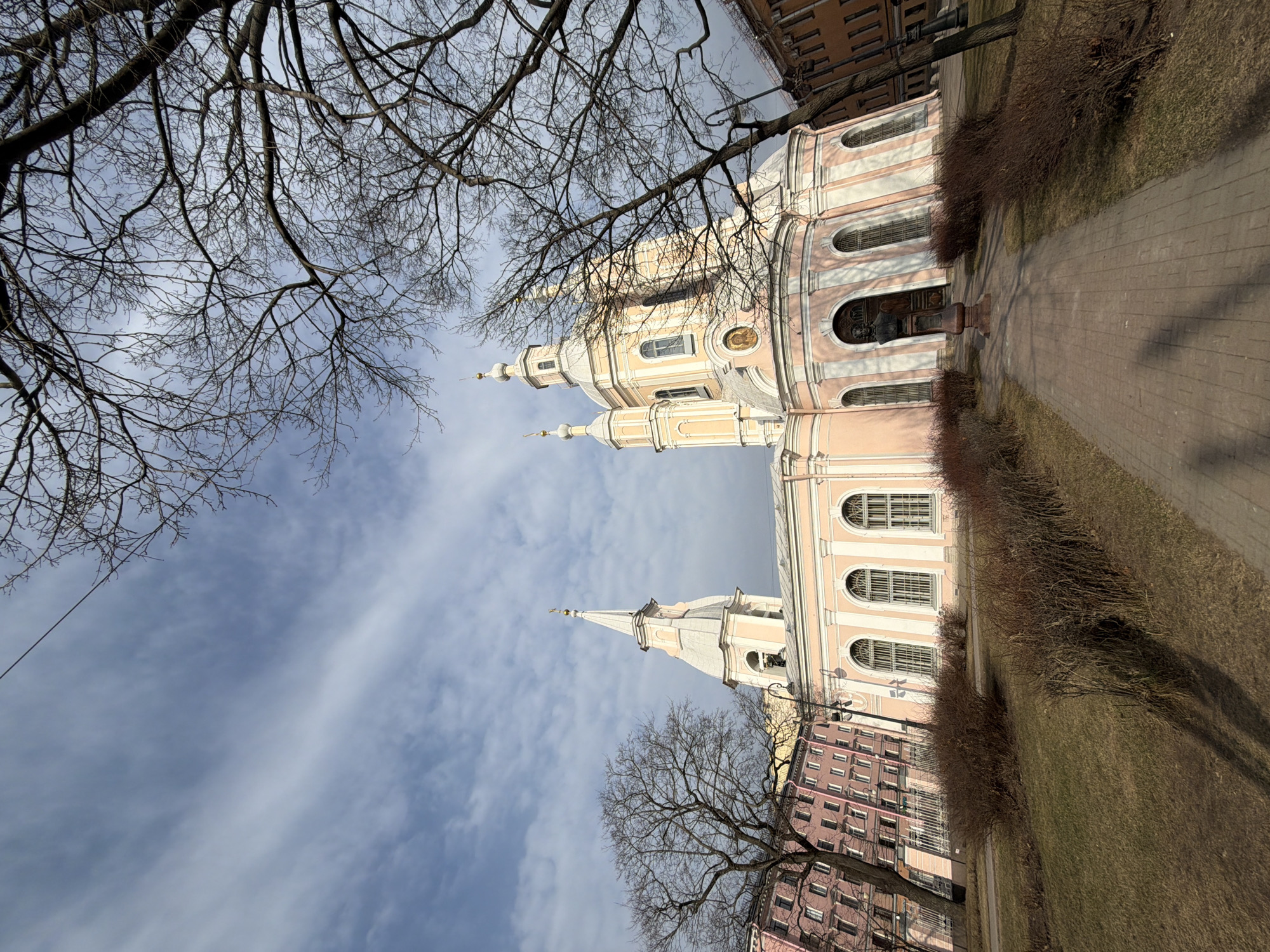 The image shows the Smolny Cathedral in Saint Petersburg, Russia. The cathedral, painted in light pink and white, is viewed from a low angle, emphasizing its height and architectural details such as the domes, spires, arched windows, and decorative elements. The cathedral is flanked by buildings, one on the left with a similar color scheme and design to the cathedral itself, and another on the right which appears to be a residential building with a darker, brick-like facade. A paved walkway and grassy area are visible on the right side of the cathedral, with bare bushes lining the walkway. In the foreground, bare tree branches frame the upper portion of the image, contrasting with the blue sky filled with scattered clouds. The lighting suggests it is daytime, with soft shadows visible on the buildings.