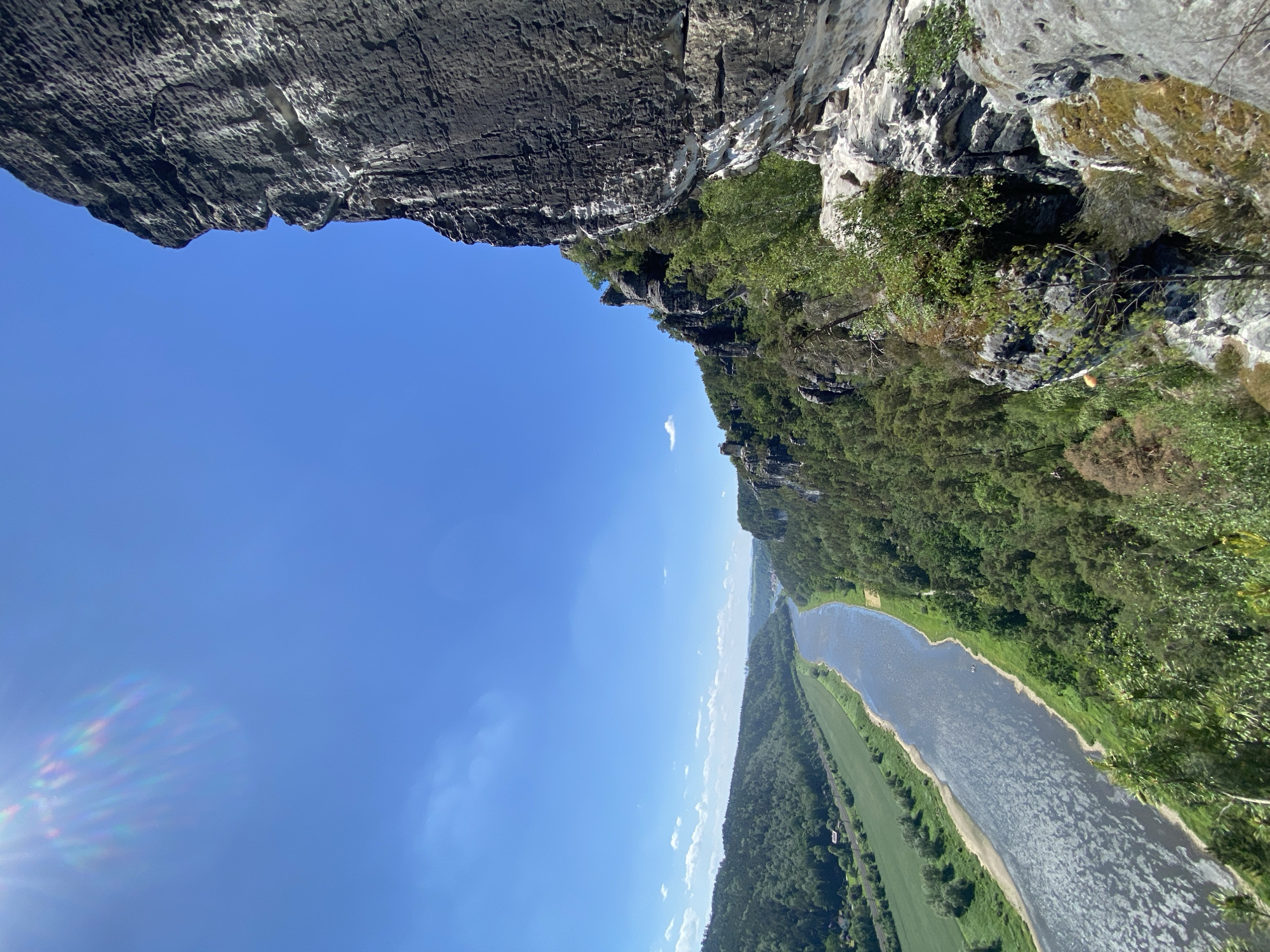 View from Bastei rock formation in Lohmen, Germany, showing the Elbe River winding through a forested valley on a sunny day. The sandstone cliffs of the Bastei are visible in the upper portion of the frame, contrasting with the blue sky and the green landscape below.