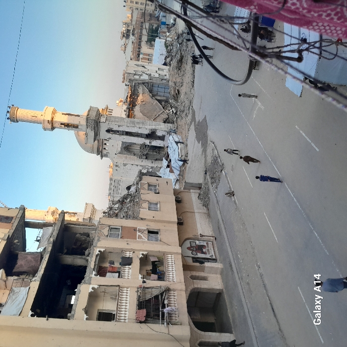 An elevated viewpoint captures a street scene in Khan Yunis, Palestinian Territory, marked by extensive structural damage. In the mid-ground, the heavily damaged Great Mosque of Khan Yunis features a partially intact minaret and a collapsed main structure, exposing its interior and generating large piles of rubble. To the left, a multi-story residential and commercial building also shows significant destruction, with upper floors partially collapsed and interiors exposed; lower balconies remain, some with laundry hanging. The ground floor displays a faded commercial sign for "NADER FASHION".
The paved street in the foreground and mid-ground has white lane markings and is mostly clear of large debris, though smaller rubble lines its edges. Approximately ten individuals, primarily males, are observed on the street; some walk in the mid-ground, one sits on a curb near the damaged building, and others gather near the mosque's rubble. The scene is illuminated by soft, warm light consistent with late afternoon or early morning, under a clear sky. Visible in the lower-left corner is a "Galaxy A14" watermark.