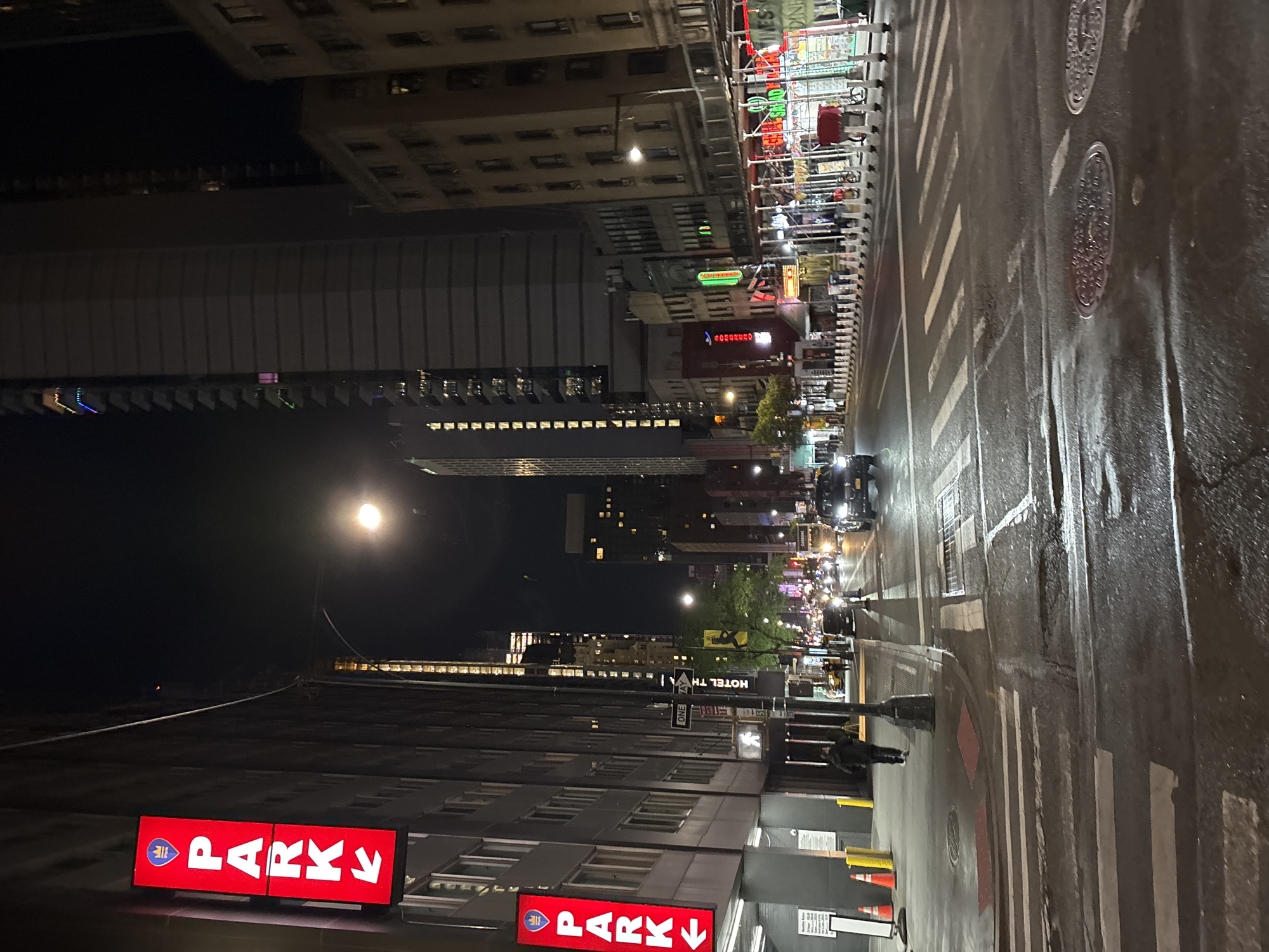 This image captures a wide-angle view of a city street at night, likely after a recent rain, given the wet and reflective pavement. Tall, multi-story buildings line both sides of the street, many with illuminated windows, contributing to the urban glow.
The scene is bustling with typical nighttime city activity. Cars with illuminated headlights can be seen on the wet asphalt road, which features prominent white crosswalk markings and several manhole covers. Numerous artificial light sources, including streetlights, building lights, and colorful neon business signs, brightly illuminate the scene and reflect off the wet ground. One pedestrian is visible walking on the sidewalk towards the lower right.
Notable details include a bright street lamp on the left, traffic cones on the sidewalk, and various signs. Visible text includes two prominent red signs with white letters reading "PARK," each featuring an arrow (one pointing down, one pointing right) and a small blue logo with a white icon. A white "ONE WAY" sign is visible in the middle distance. Other partially discernible text from neon signs includes fragments that appear to be "HOTEL TH..." (likely "Hotel The..."), hints of "SALAD," "Gourmet," "Diner," and a blue sign that might read "DOMINO" or similar. The overall ambiance is that of a vibrant, active metropolitan area at night, characteristic of New York.