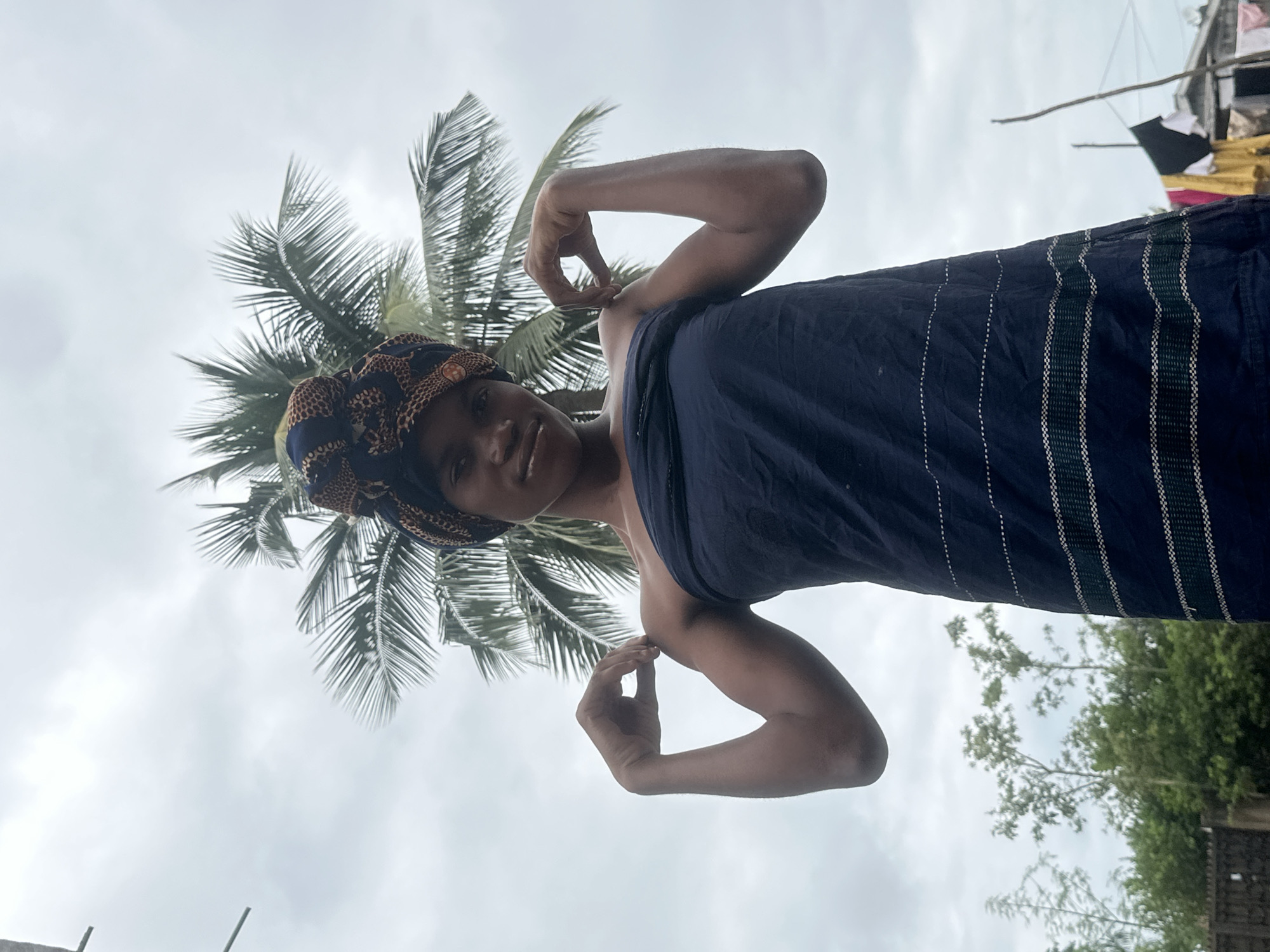 An upward-angled, medium shot captures a young Black woman standing outdoors against a cloudy sky and a palm tree. She is wearing a dark blue strapless dress and a patterned headwrap. Her right arm is bent at the elbow, with her hand resting on her shoulder, while her left arm is also bent, with her hand positioned near her shoulder, as if flexing her bicep. The background shows lush green trees and foliage, and in the far right, laundry hangs on a clothesline near some buildings. The scene appears to be in a residential area.