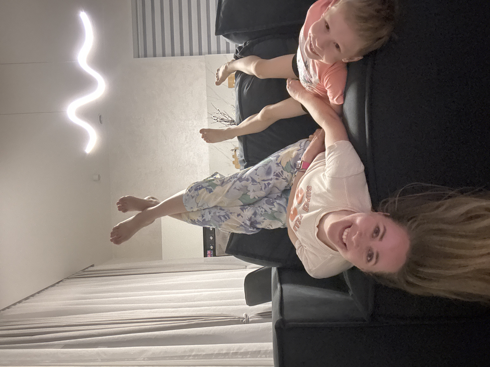 A woman and a child are lying on a dark sofa in a room. The woman is in the foreground, smiling at the camera with her head near the bottom of the frame and her long blonde hair spread out on the sofa. She is wearing a white t-shirt and floral patterned pants. The child is lying on the back of the sofa near the top of the frame, wearing a light pink shirt. Both are barefoot. In the background, there are striped curtains, a white wall, and a modern, wavy light fixture. There is also a small decorative object on a shelf. The location is the city of Chaikovsky, Russia.