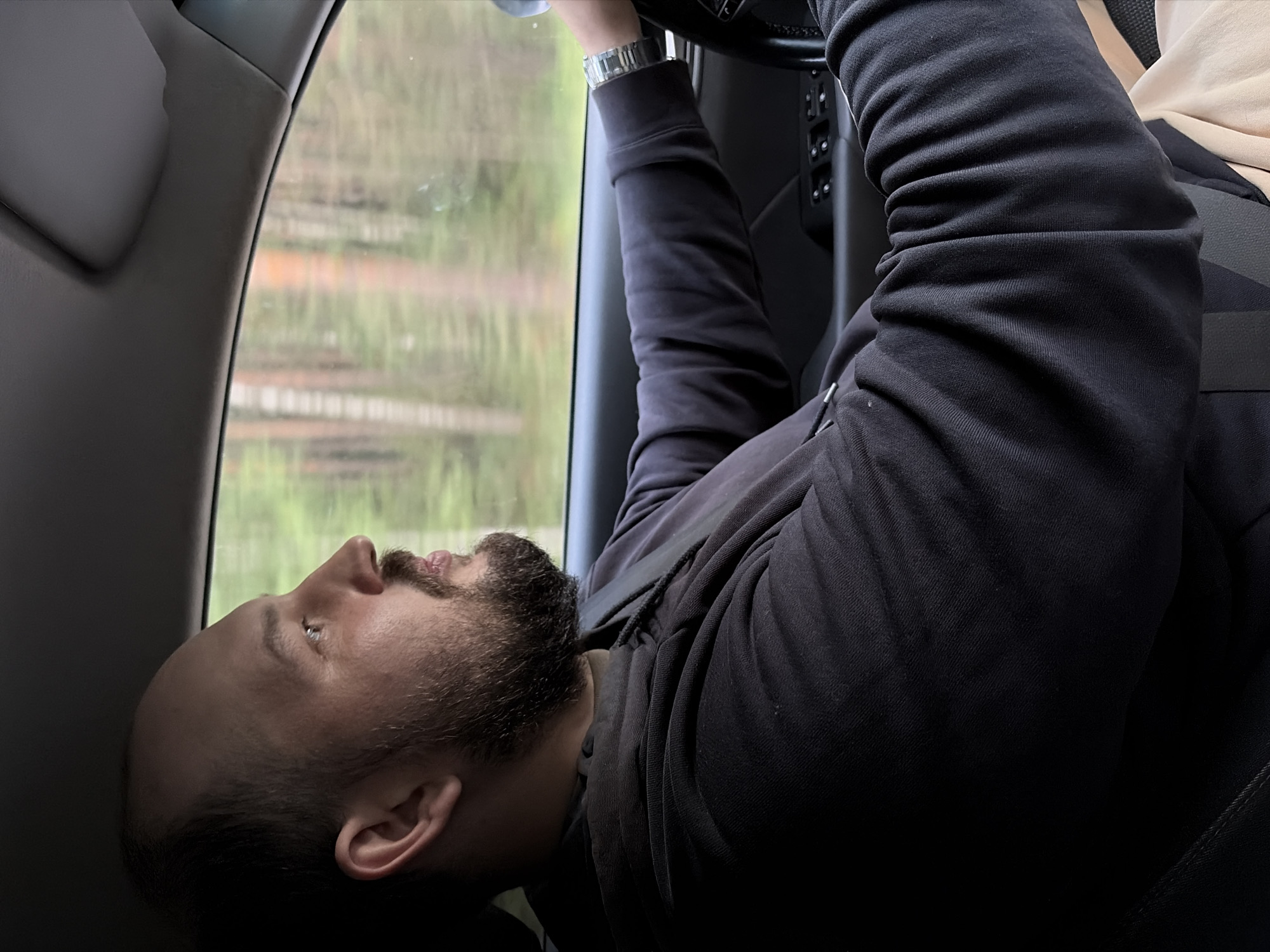 A medium shot shows a man with a beard and a shaved head driving a car, seen from the side. He is looking straight ahead through the windshield, his hands on the steering wheel. He is wearing a dark hooded sweatshirt and a seatbelt. The background outside the car window shows a blurred view of trees, suggesting a rural or forested area. The car interior is dark. The man's facial expression is neutral.
