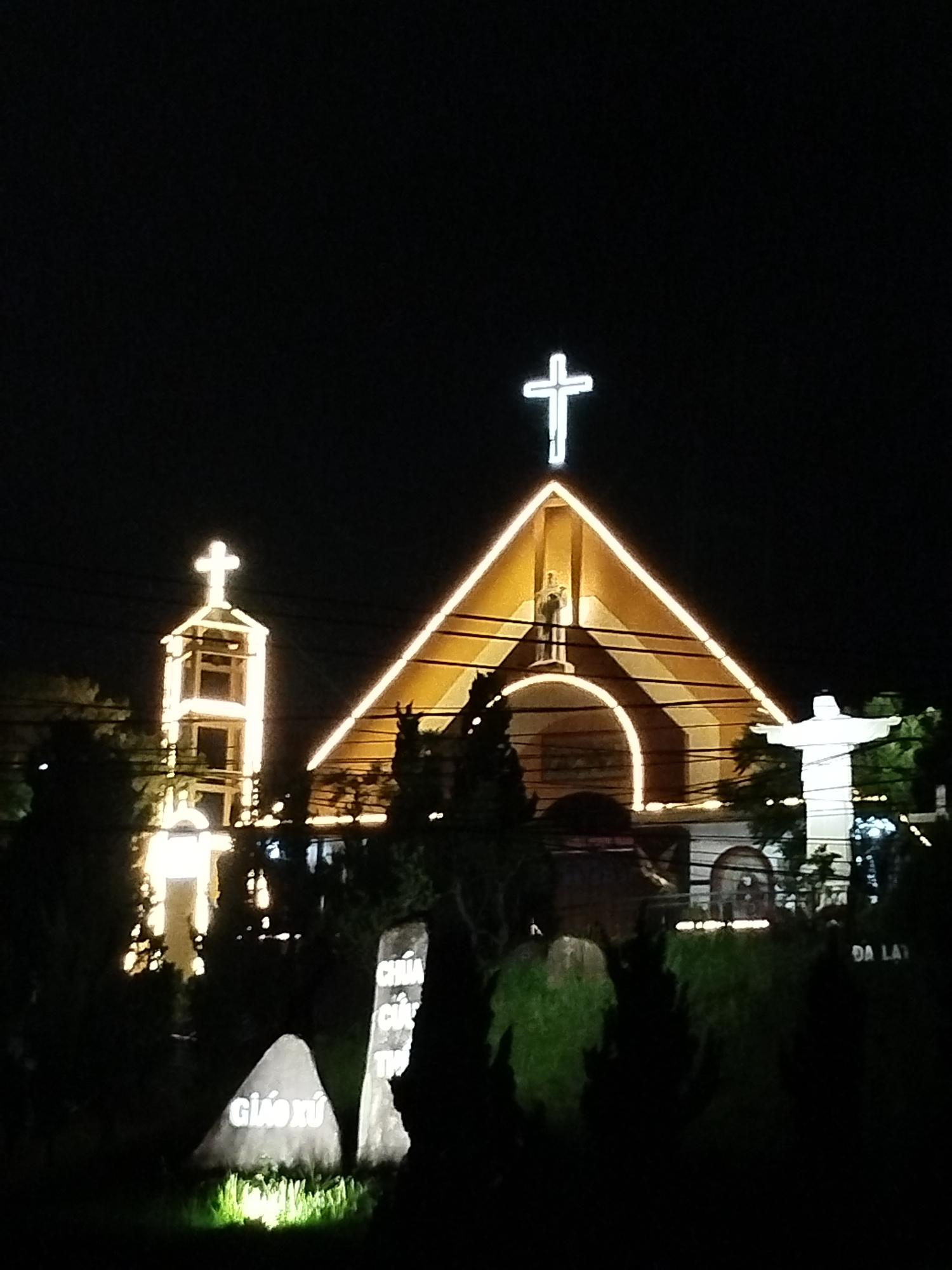 The image depicts a church at night, likely in Lạc Dương, Vietnam. The church has a large, triangular facade illuminated with lights outlining its shape and a cross at the peak. A smaller bell tower is also lit up with a cross on top. A statue of Jesus is on the right, also illuminated. In the foreground, there are tombstones. One reads "Giáo Xứ." The background is dark, suggesting it is night. The scene appears calm and serene.