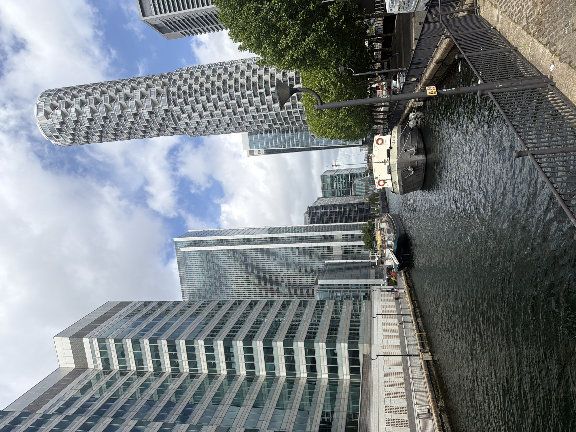 The image shows a canal in London with several high-rise buildings in the background. The buildings have glass facades and appear to be modern office or residential structures. Two boats are docked along the canal. A metal railing and a paved walkway run along the edge of the canal. The sky is partly cloudy. The image is likely taken in the Canary Wharf area of London.