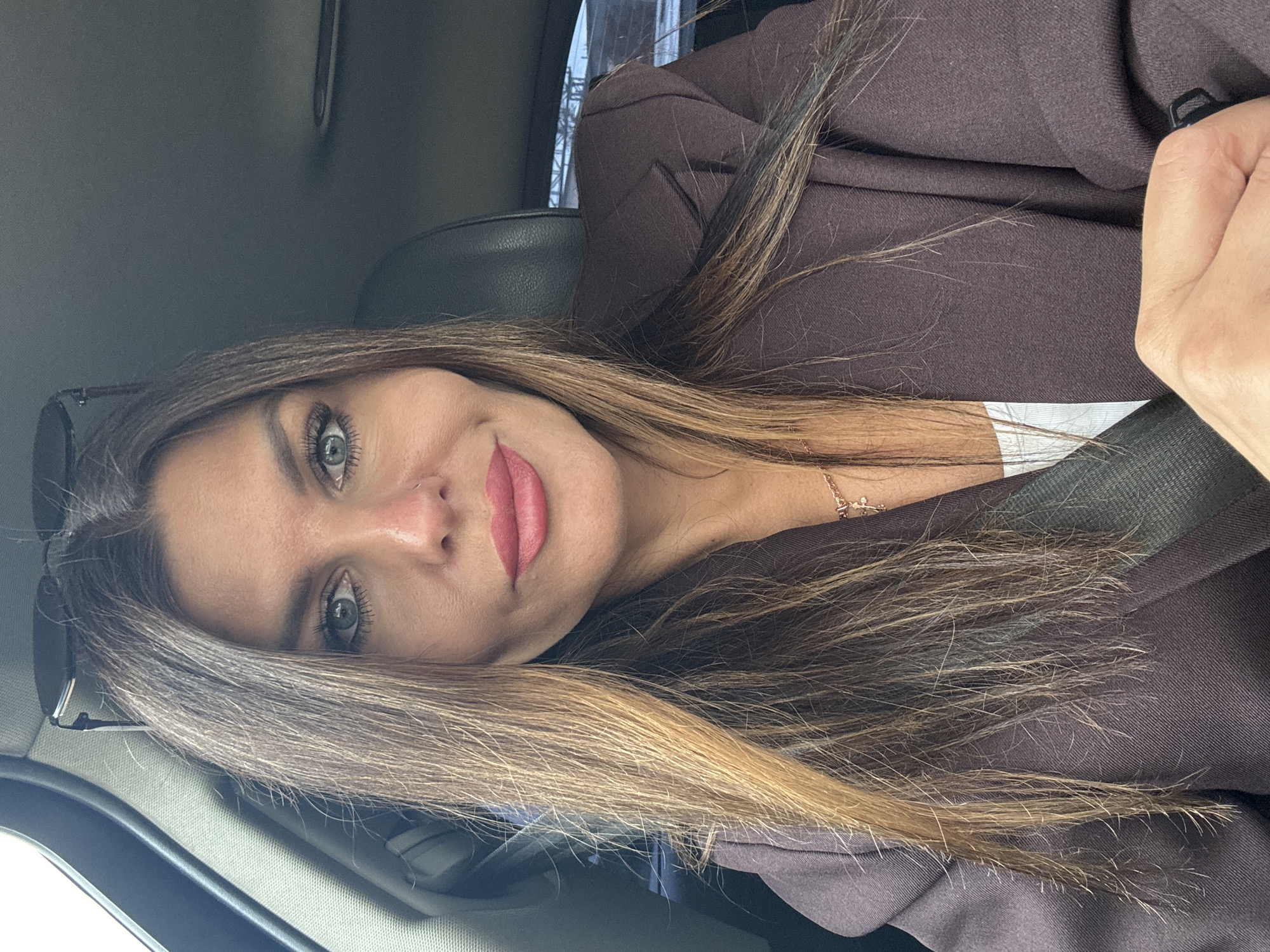 A woman with long brown hair and blue eyes is sitting in a car. She is wearing a brown blazer and a white top. She is looking at the camera and smiling. The car appears to be a personal vehicle, possibly a sedan, and is in an urban environment. The lighting suggests it is daytime. There is no specific information to place this image in Chaikovsky, Russia, as the background is generic.