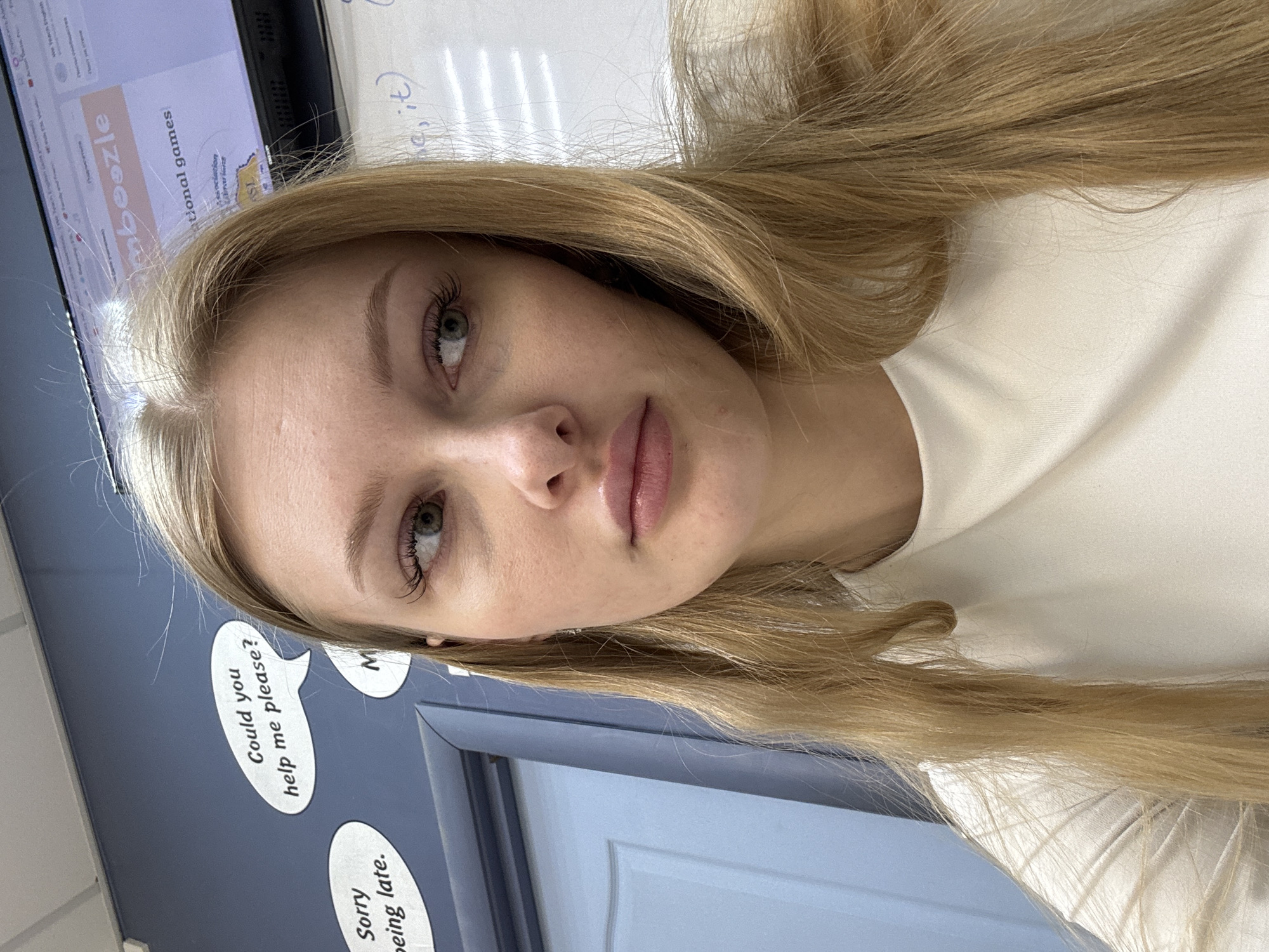A close-up, portrait-style photograph features a fair-skinned woman with blonde hair. She has light eyebrows and long eyelashes, and her lips appear full and slightly pink. The woman is wearing a white top, visible on the right side of the image. The background is varied. To the left, there is a blue wall with two speech bubble-shaped signs that contain the text "Could you help me please?" and "Sorry being late." Above and to the left is a monitor displaying the word "mboozle", "national games", "Association" as well as some additional text that is not legible. To the right of the woman is a white board with illegible writing. The city of Perm, Russia, is not discernible from this image.