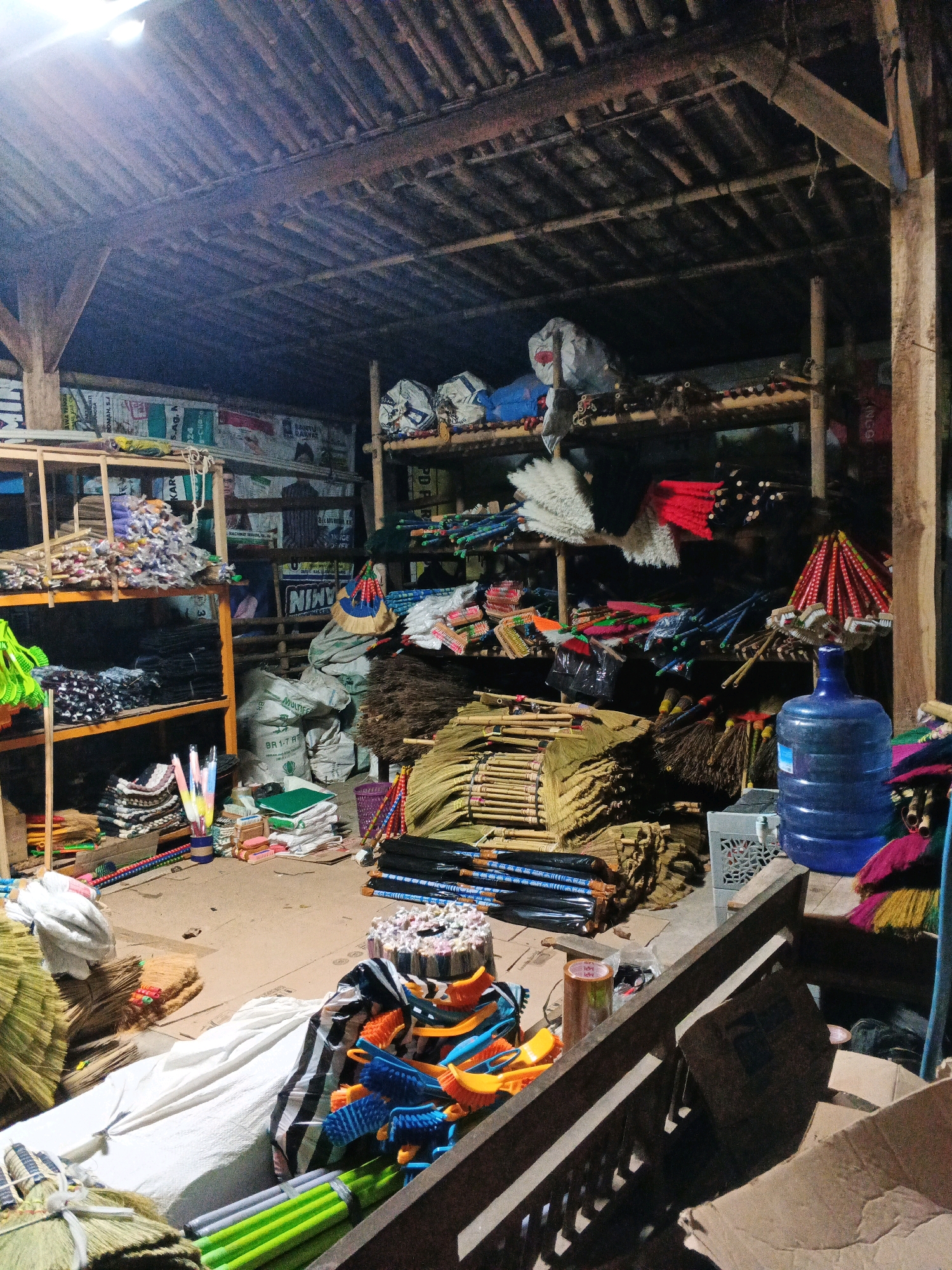 The image depicts the interior of a shop in Sirau, Indonesia, filled with a variety of cleaning supplies, primarily brooms and brushes. The shop appears to be somewhat disorganized, with items stacked on shelves, piled on the floor, and leaning against structures.
In the foreground, on the left, a stack of green-handled brushes is visible, alongside what appears to be fabric or cleaning cloths. Further back, a large pile of brooms with natural fibers is arranged. In the center of the image, there are several stacks of brooms with wooden handles and bundles of brushes with colorful plastic handles. A white sheet covers a large object on the floor. To the right, a large blue water jug is prominent, with a white plastic crate next to it. Various bags, some appearing to be filled with materials, are present throughout the space.
Shelving units, constructed from wood and possibly bamboo, are used to store more items. These shelves hold stacks of what look like smaller brooms or brushes with colorful bristles, as well as what might be spare parts or materials for cleaning tools. The walls behind the shelves are adorned with what seem to be sacks or posters with text.
The ceiling is constructed from bamboo or wooden poles and slats, characteristic of some traditional Indonesian architecture. The lighting appears to be artificial, likely from overhead lamps, casting shadows and creating a somewhat dim atmosphere. No people are visible in the image. The overall impression is that of a storage area or a small retail space for cleaning implements.