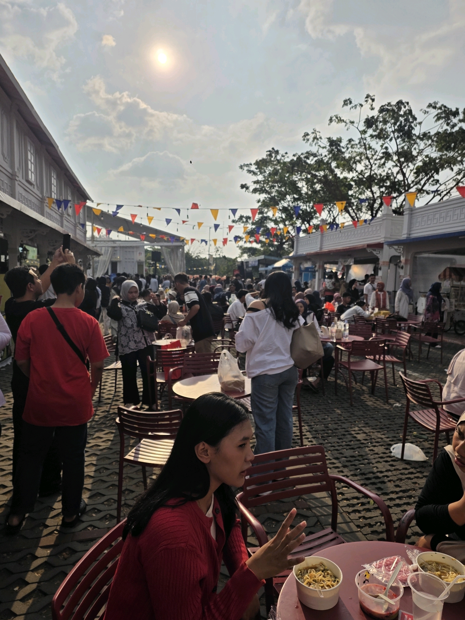 This image depicts an outdoor gathering, likely a food festival or market, in Bandung City, Indonesia, given the context provided. The scene is bustling with people of various ages, many of whom are casually dressed. Some are seated at tables, some are standing, and others are walking through the area. Several individuals appear to be eating or ordering food, with bowls of noodles and drinks visible on the tables.
The environment features a paved ground and a street lined with buildings that appear to house stalls or shops. Colorful triangular flags are strung across the area, adding a festive atmosphere. The sky is partly cloudy, with the sun visible and casting shadows on the ground. The overall impression is one of a lively daytime event.
Specifically, there are groups of people interacting, with some engaged in conversation, others taking pictures with their phones, and some focused on their food. The lighting suggests it is daytime, possibly late afternoon given the angle of the sun and the shadows. The architectural style of the buildings and the presence of food stalls are consistent with common market or festival setups in urban Indonesian settings.