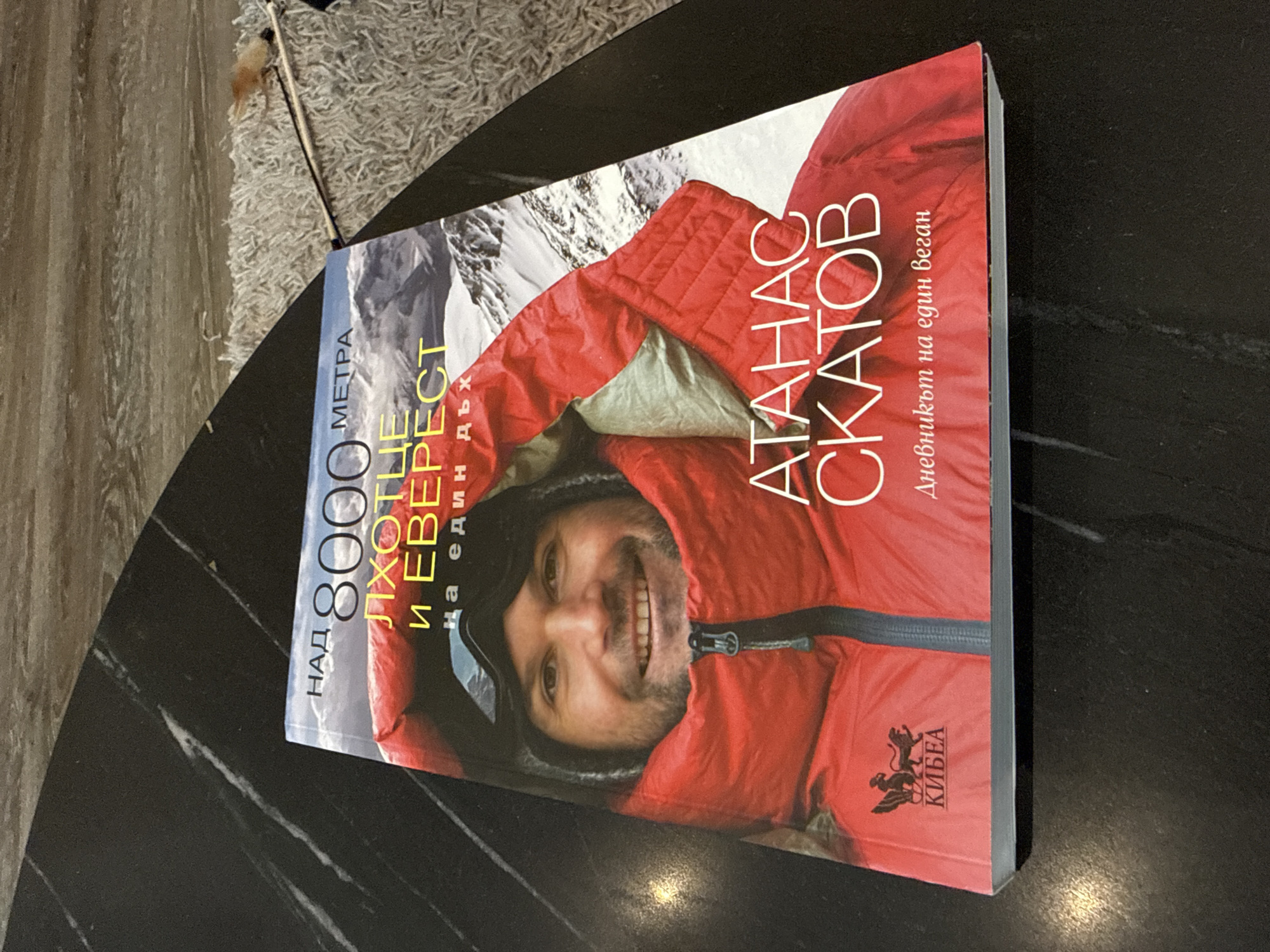 The image shows a book titled "Над 8000 Метра Лхотце и Еверест" (Above 8000 Meters Lhotse and Everest), written by Атанас Скатов (Atanas Skatov), a Bulgarian mountaineer. The subtitle translates to "Diary of a Vegan." The book cover features a photo of Skatov in mountaineering gear against a backdrop of snowy mountains. The publisher's logo, "КИБЕА" (KIBEA), is visible on the bottom right of the book cover. The book is resting on a black, marble-patterned surface. The background also includes a glimpse of a textured, light-colored rug. There is no information provided in the image to place it in Varna, Bulgaria.