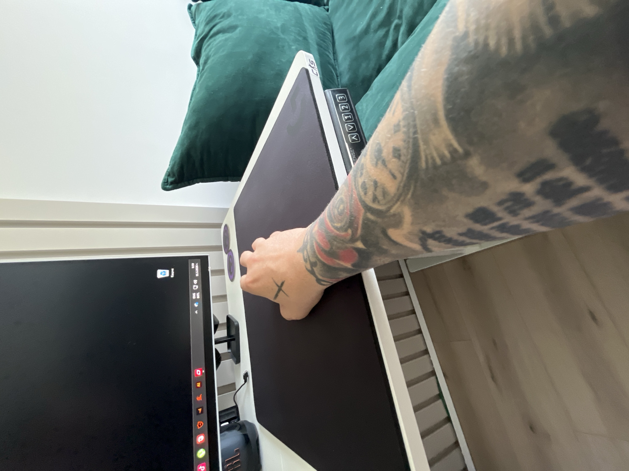 The image shows a person's tattooed arm and hand reaching towards a black surface, possibly a graphics tablet or monitor, with a white frame. The device is positioned next to a computer monitor displaying a partially visible desktop. Behind the device, there's a green velvet pillow or cushion. The flooring appears to be light-colored wood. The location, Varna, Bulgaria, is not discernible from the visual information provided.