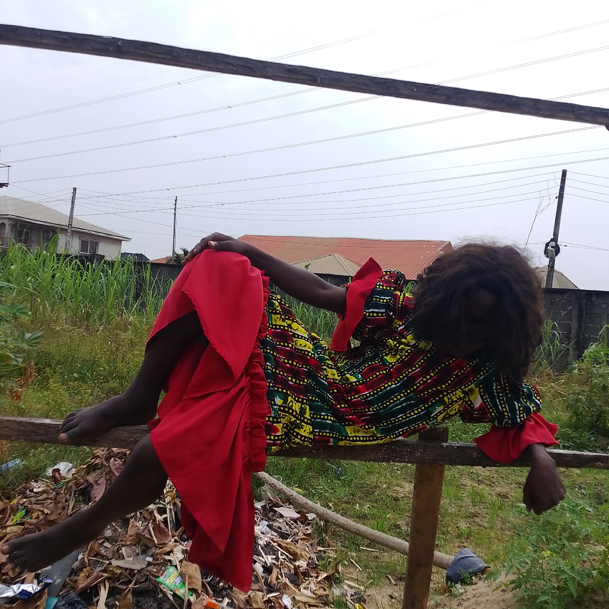 A full-body, outdoor shot depicts a dark-skinned person with dark, curly hair lying on a wooden beam. The person is wearing a red dress with a multicolored pattern of yellow, green, and black stripes. The dress has ruffled sleeves and a long, flowing skirt. The person's left arm is bent at the elbow and resting on their left leg, while their right arm is extended and bent at the elbow, with their hand resting on the beam. Their legs are also bent and their feet are resting on the beam. The person is lying on a beam above a pile of debris that includes leaves, plastic bags, and other trash. Behind the person is a dense green area of grass and tall stalks of vegetation, with some houses and electrical poles visible in the distance. The sky is overcast and gray. The scene appears to be located in Ado, Nigeria.