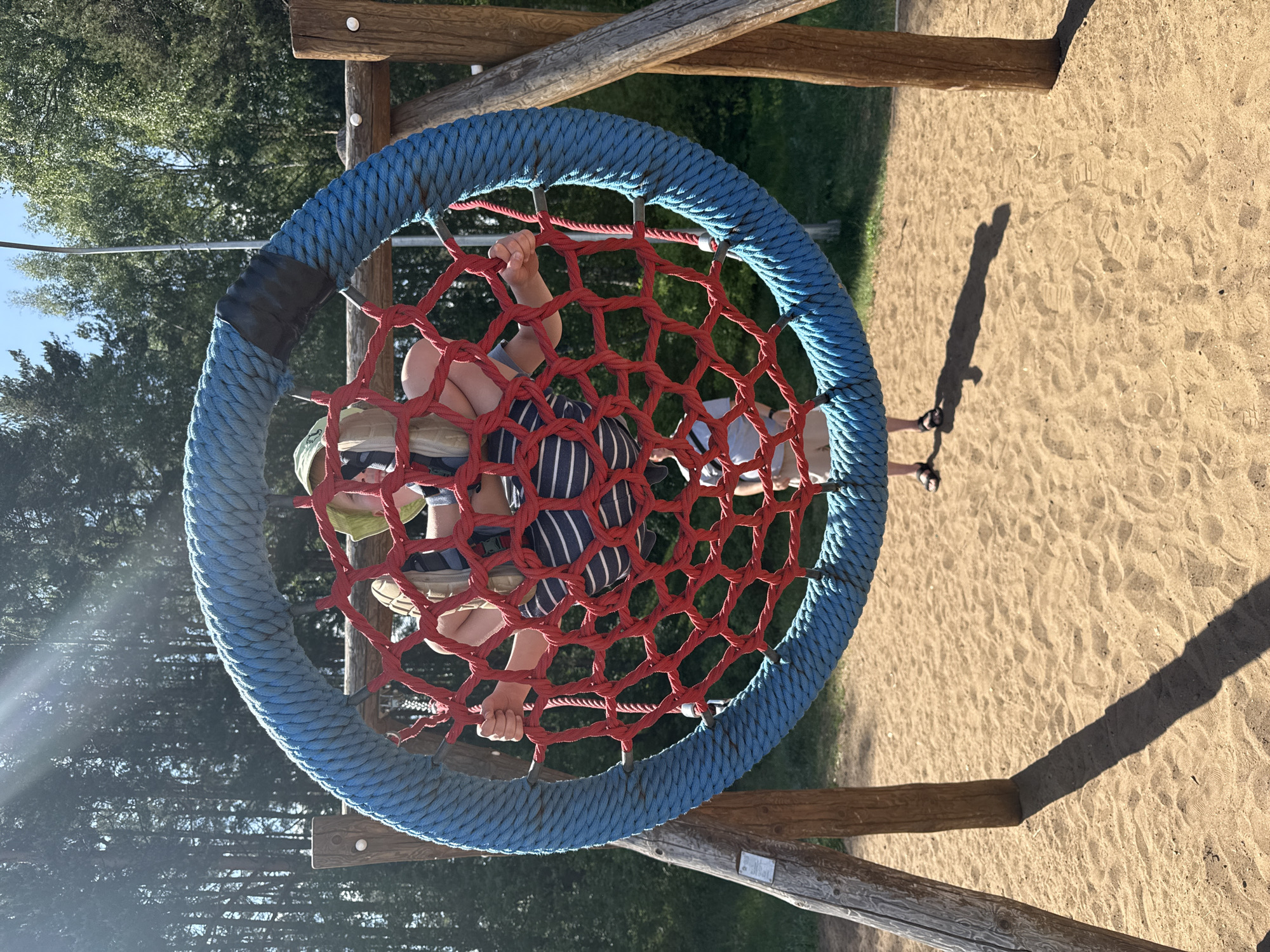 A child wearing a green hat and striped clothing is inside a circular net swing at a playground. The swing has a blue braided border and a red net. The playground has a sandy surface. The photograph was taken in Chaikovsky, Russia.