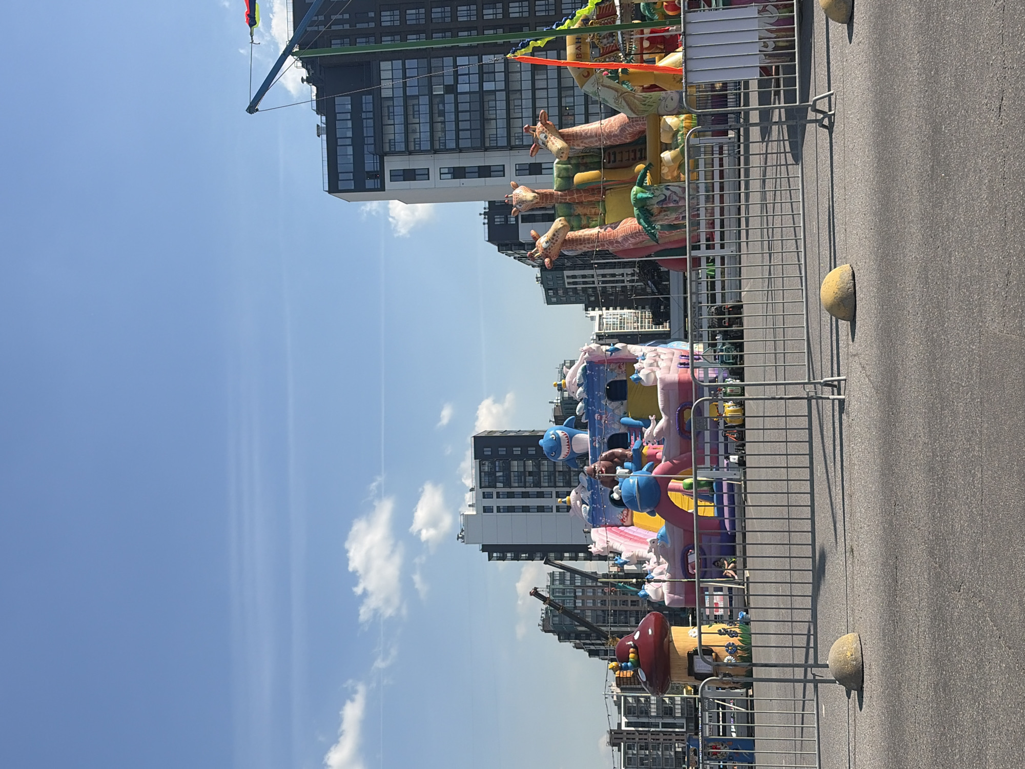 This image captures an outdoor scene in Saint Petersburg, Russia, likely a public space set up for entertainment. In the foreground, metal barricades with concrete bases line a paved surface, possibly a street or parking lot. Beyond the barricades, a vibrant inflatable playground is the focal point. This structure features various cartoonish elements, including a large shark, a mushroom-shaped playhouse, and a cluster of giraffes. Inflatable slides and climbing areas are also visible. The playground is surrounded by additional fencing and some carnival-style elements like colorful flags and signage.
In the background, the urban landscape of Saint Petersburg is evident. Modern residential buildings with dark-paned windows and balconies rise into the clear blue sky. Construction cranes are also present, suggesting ongoing development in the area. The sky is bright with scattered white clouds. The overall impression is one of a sunny day in an urban environment, with a dedicated area for children's entertainment. No people are clearly visible engaged in activities within the playground itself.