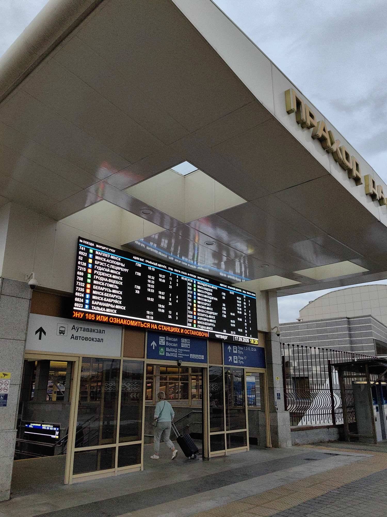 The image shows the entrance of a train station in Minsk, Belarus. A large digital display board lists train departures and destinations. In the foreground, a woman is walking with a suitcase towards the station entrance. The station appears to be modern with large glass windows and a covered walkway. The sky is overcast, suggesting a cloudy day. The overall scene depicts a typical transportation hub environment.